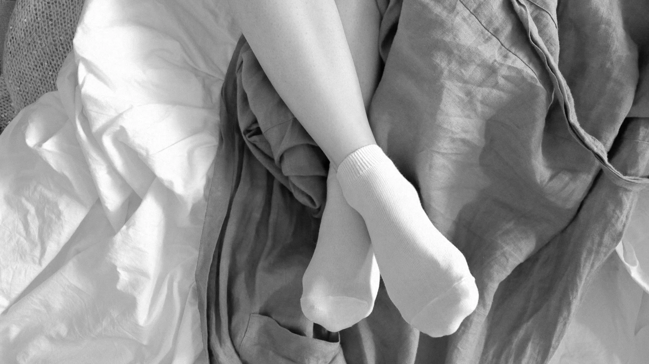 Nahaufnahme von entspannten Füssen in weissen Socken, liegend auf einer Decke oder Bettwäsche, die aus verschiedenen Stoffen besteht.