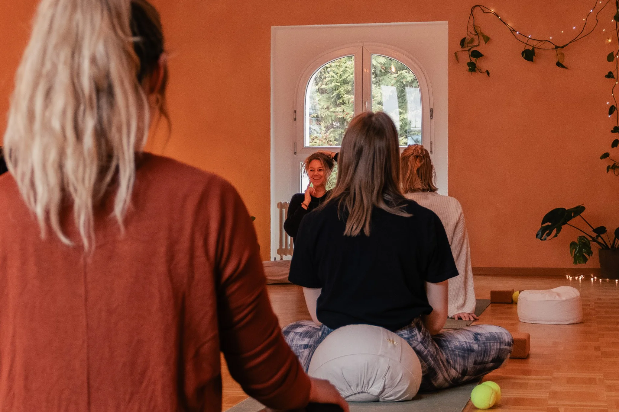 Eine Gruppe von Frauen sitzt im Schneidersitz auf Yogamatten in einem Raum mit orangefarbenen Wänden und einem großen Fenster im Hintergrund, während eine Frau in Schwarz vorne den Raum leitet und lächelt.