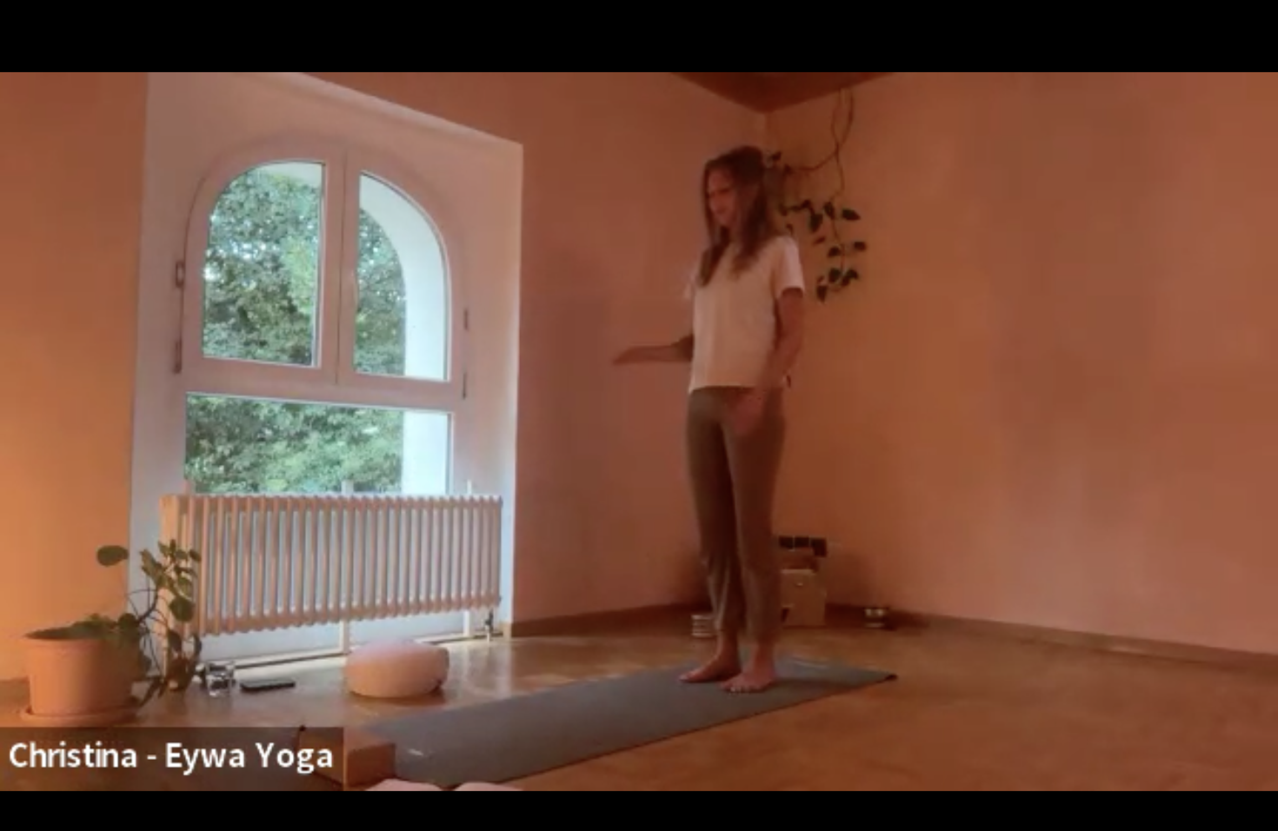 Eine Frau macht Yoga in einem hell erleuchteten Raum mit großen Fenstern, durch die Bäume sichtbar sind.
