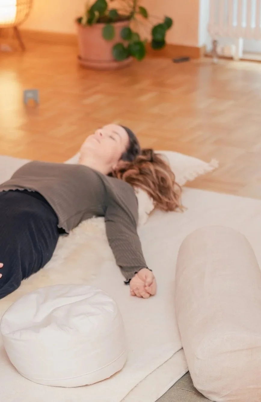 Eine Frau liegt entspannt auf einem weißen Teppich im Wohnzimmer, die Augen geschlossen, neben ihr ein Kissen und eine Decke. Thai Yoga Massage Bern.