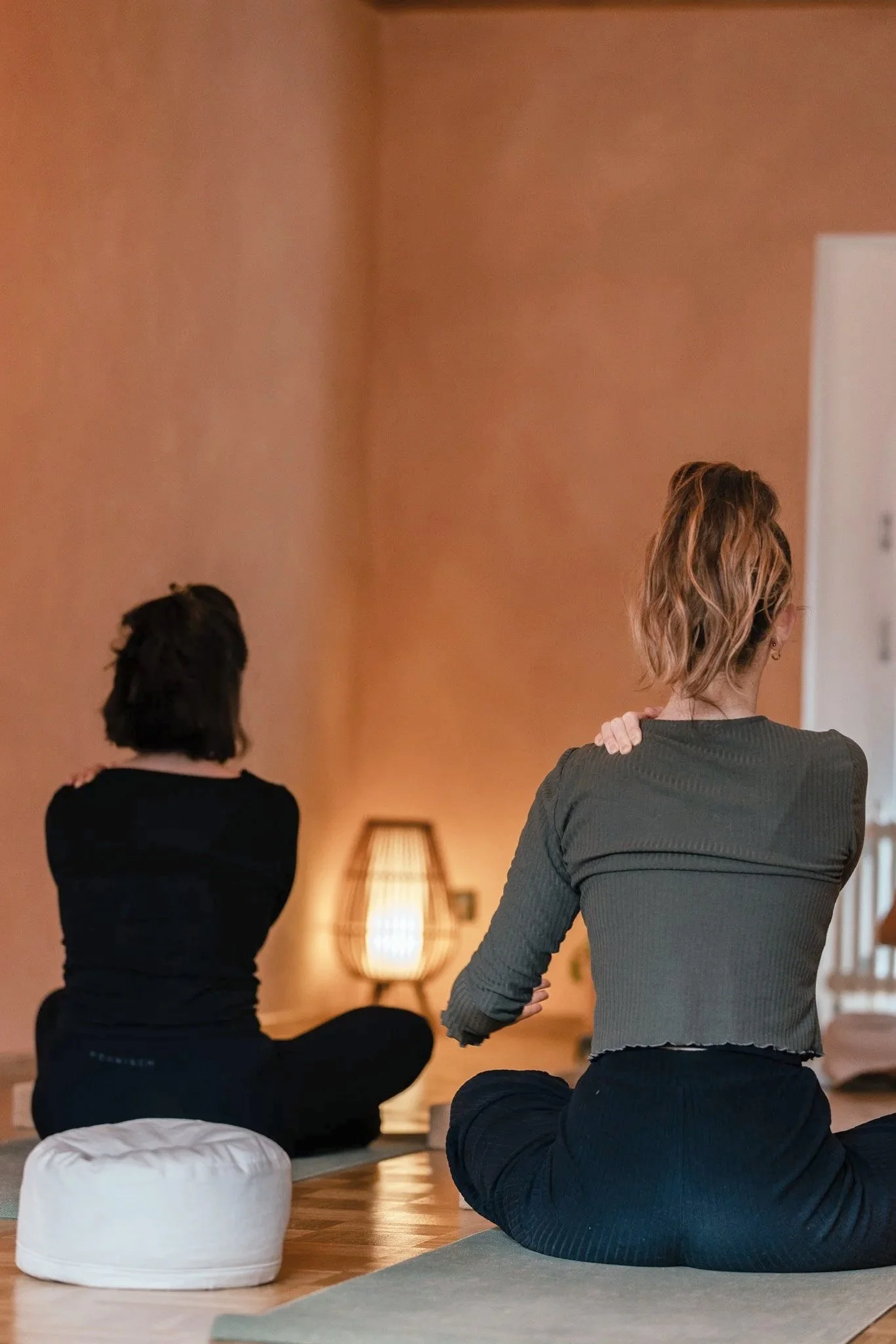 Zwei Frauen sitzen in einer Yoga-Pose auf Kissen im Wohnzimmer, während eine Yoga-Lehrerin neben ihnen steht, um ihnen Anleitung zu geben. Somatic Yoga Bern.