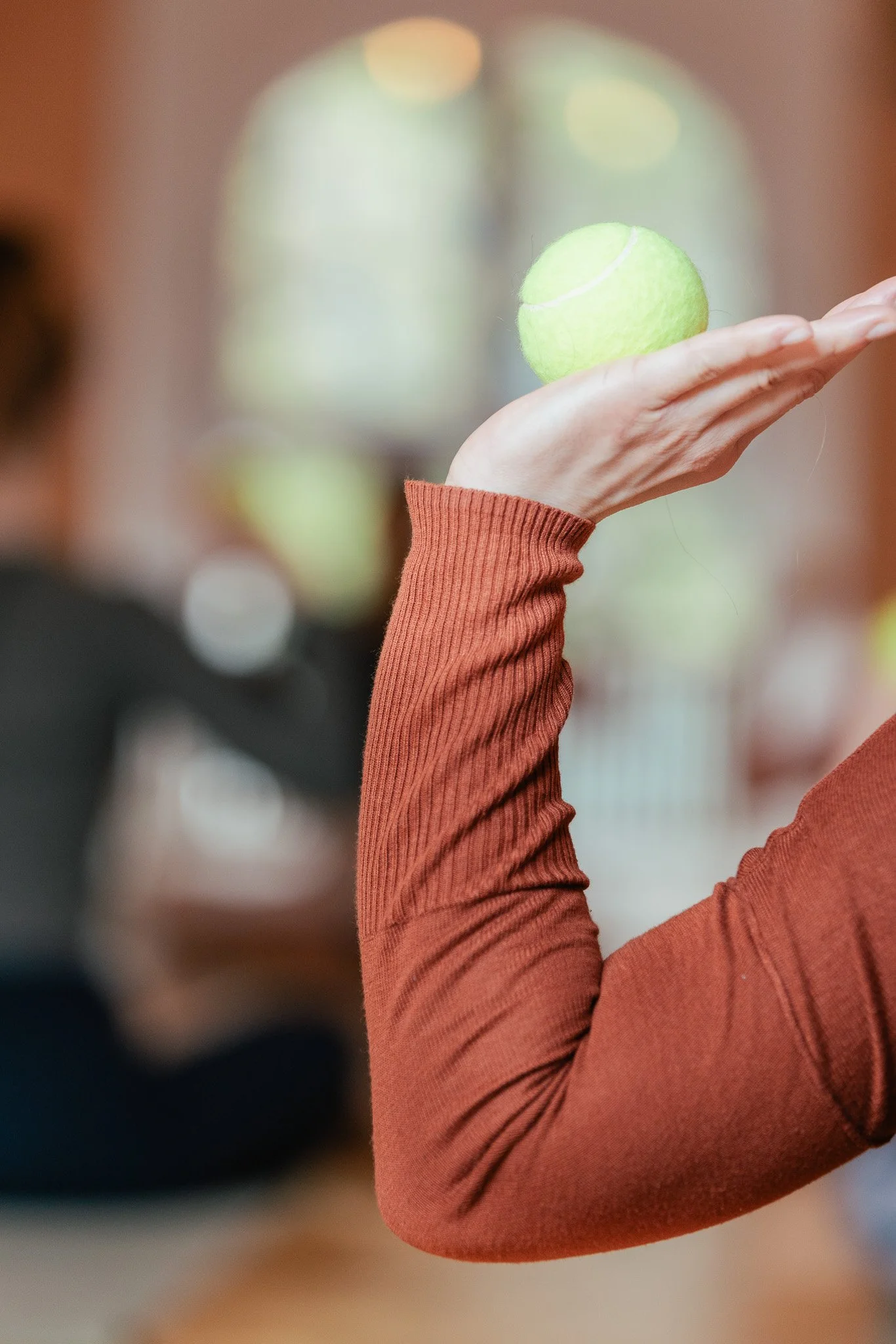 Nahaufnahme einer Person, die eine grüne Tennisball auf ihrer Hand hält, getragen von einem langärmligen, braunen Oberteil. Hintergrund unscharf, vermutlich in einer Indoor-Umgebung.