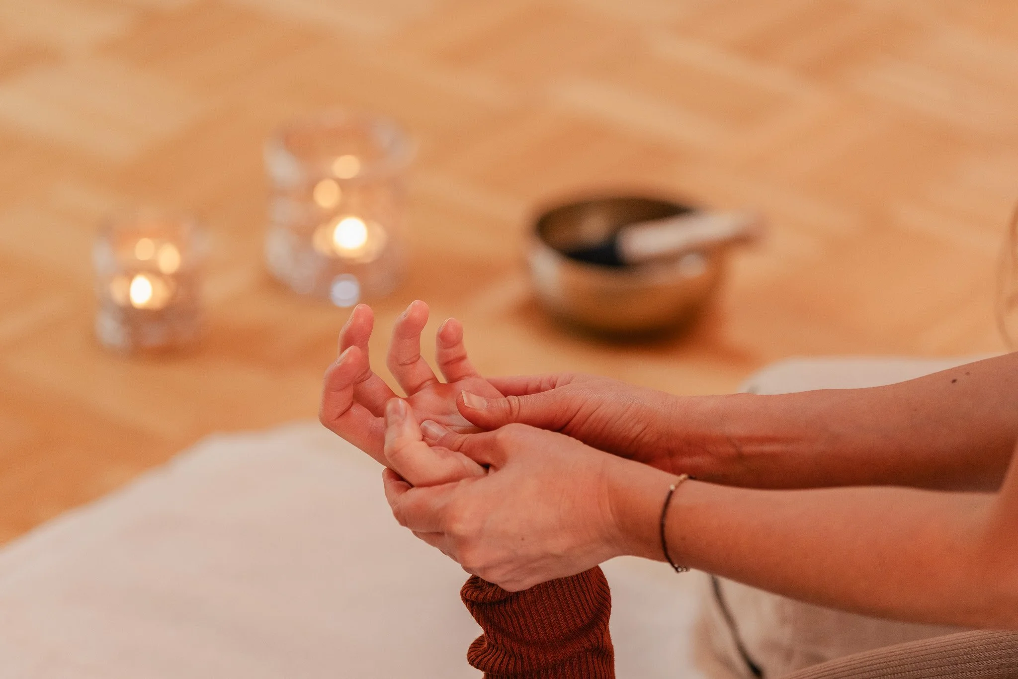 Eine Person, die eine andere Person die Hand hält, die auf ihrem Knie liegt. Im Hintergrund sind zwei Kerzen, eine Schale und ein Löffel sichtbar. Thai Yoga Massage Hände. Nervensystem. Traumasensitiv.