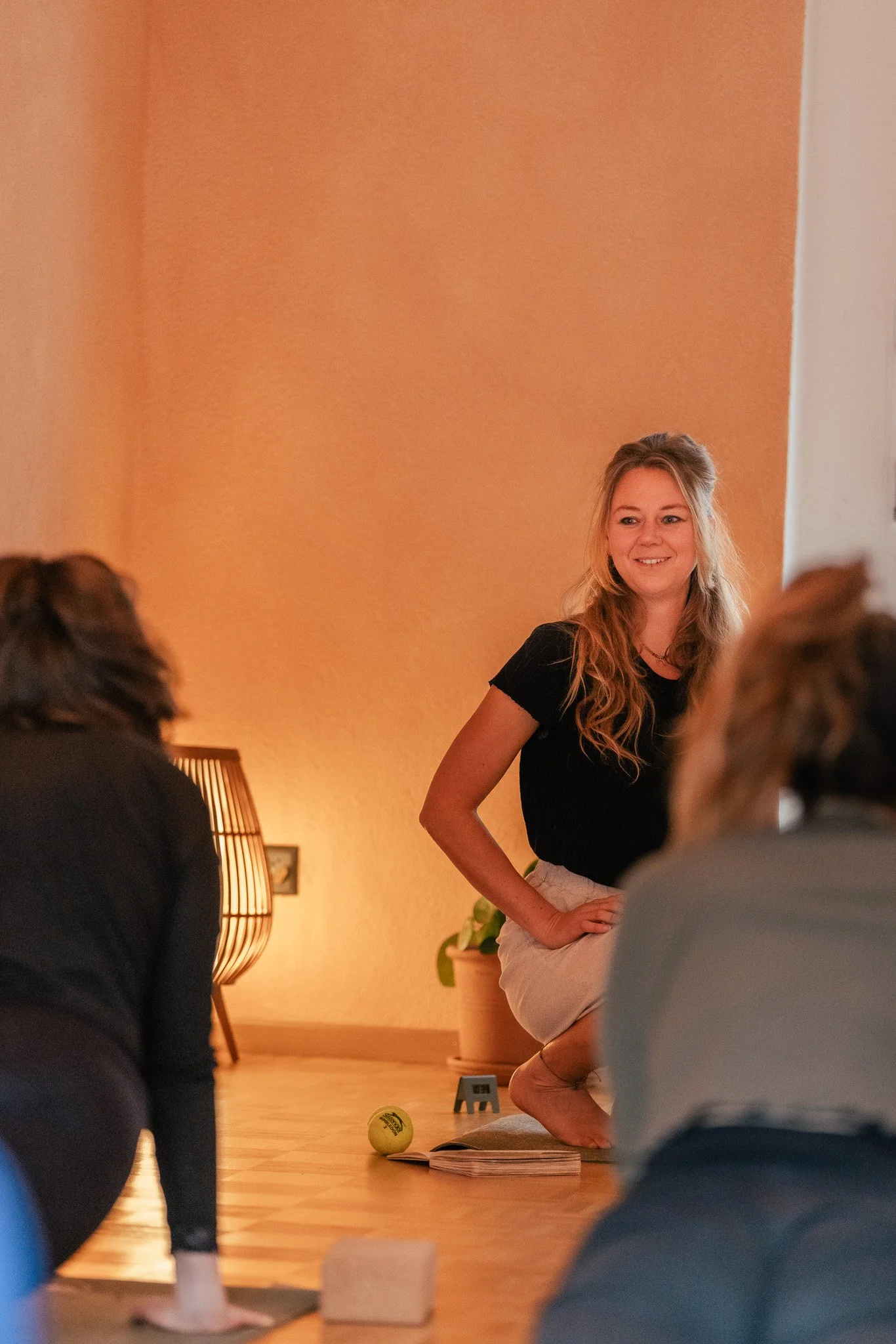 Eine Frau sitzt auf dem Boden in einer Yoga- oder Meditationshaltung, während sie in einer Gruppe von Menschen vor einem warm beleuchteten Raum spricht oder präsentiert. Somatic Yoga. Yoga. Traumasensitiv.