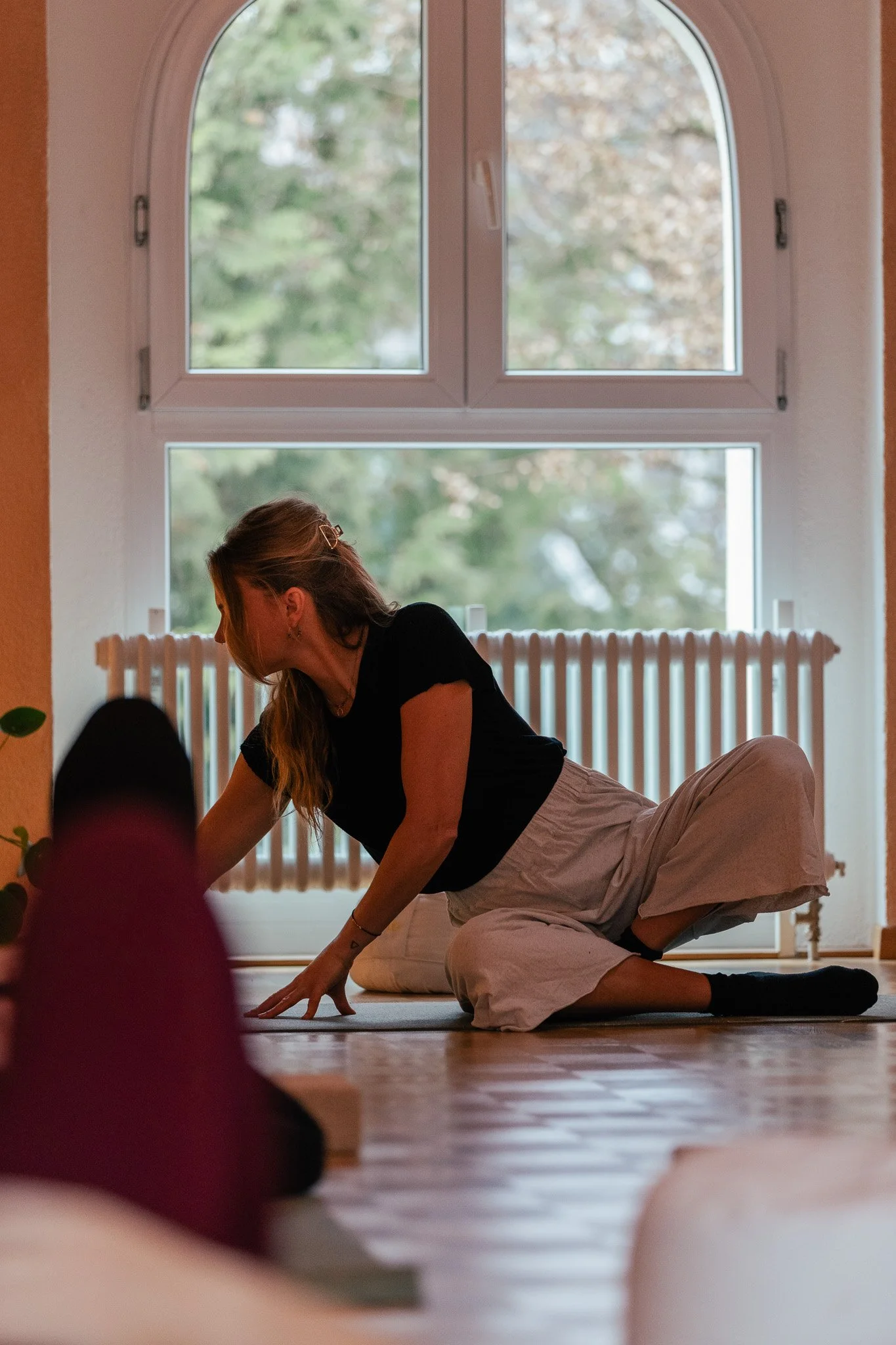 Eine Frau macht Yoga auf einem Boden vor einem großen Fenster, durch das Bäume draußen zu sehen sind.