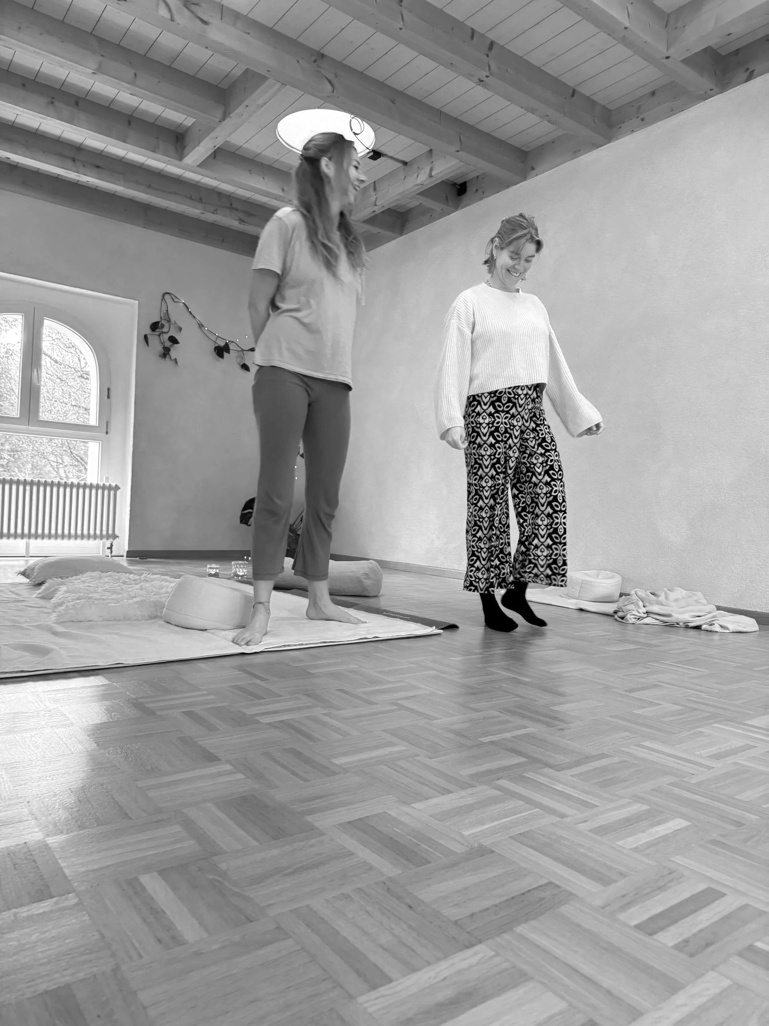 Christina Winzeler und eine Klientin bei in einem Raum mit Holzbalkendecke und Holzboden, lachend und in bequemer Kleidung, im Hintergrund eine Wand mit Dekoration. Eine Sitzung der Somatischen Prozessbegleitung, die Körperwahrnehmung fördert.