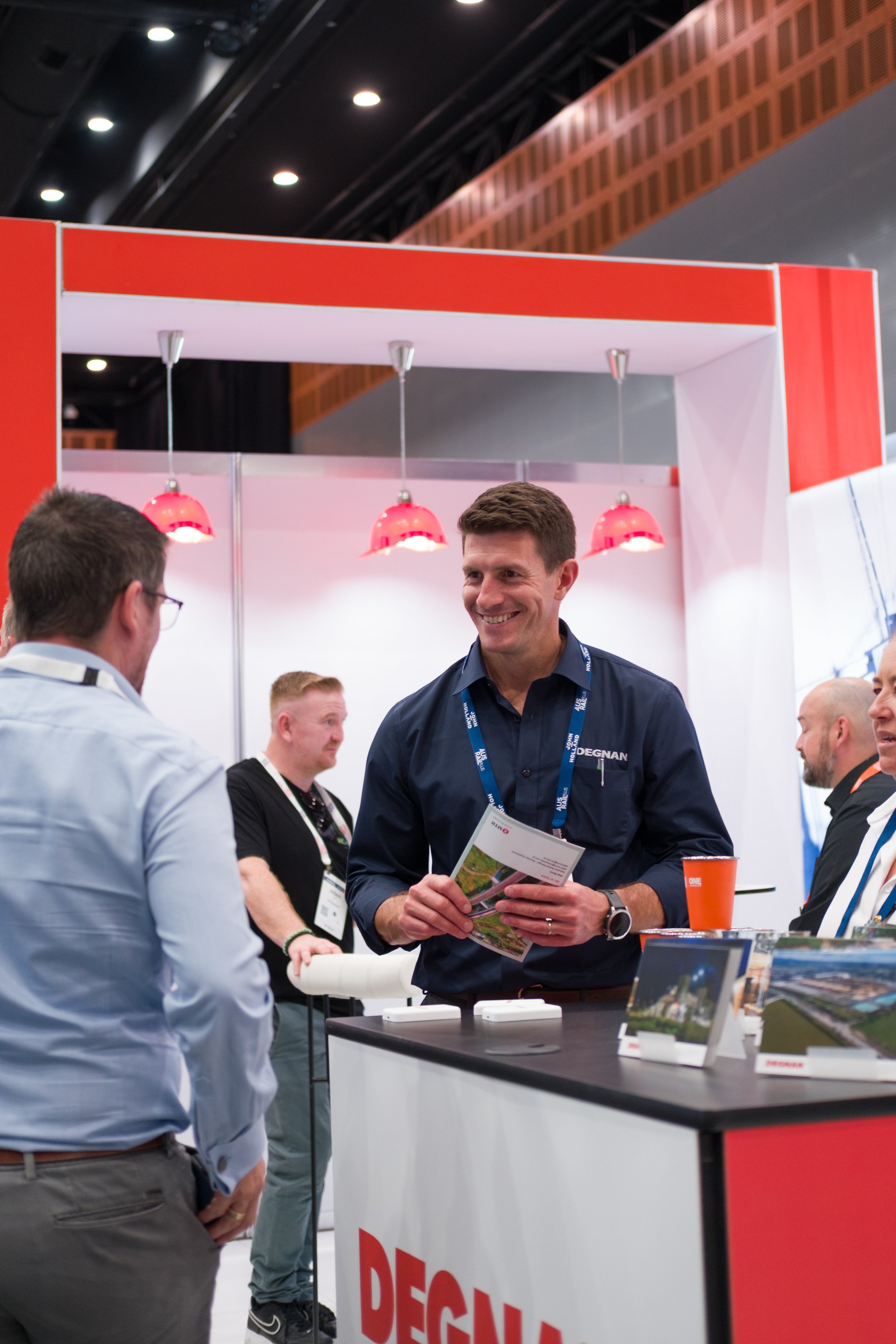 A man in a navy blue shirt talking to another man at a trade show booth, smiling and holding a brochure. Several people are in the background, and the booth has a red and white color scheme.