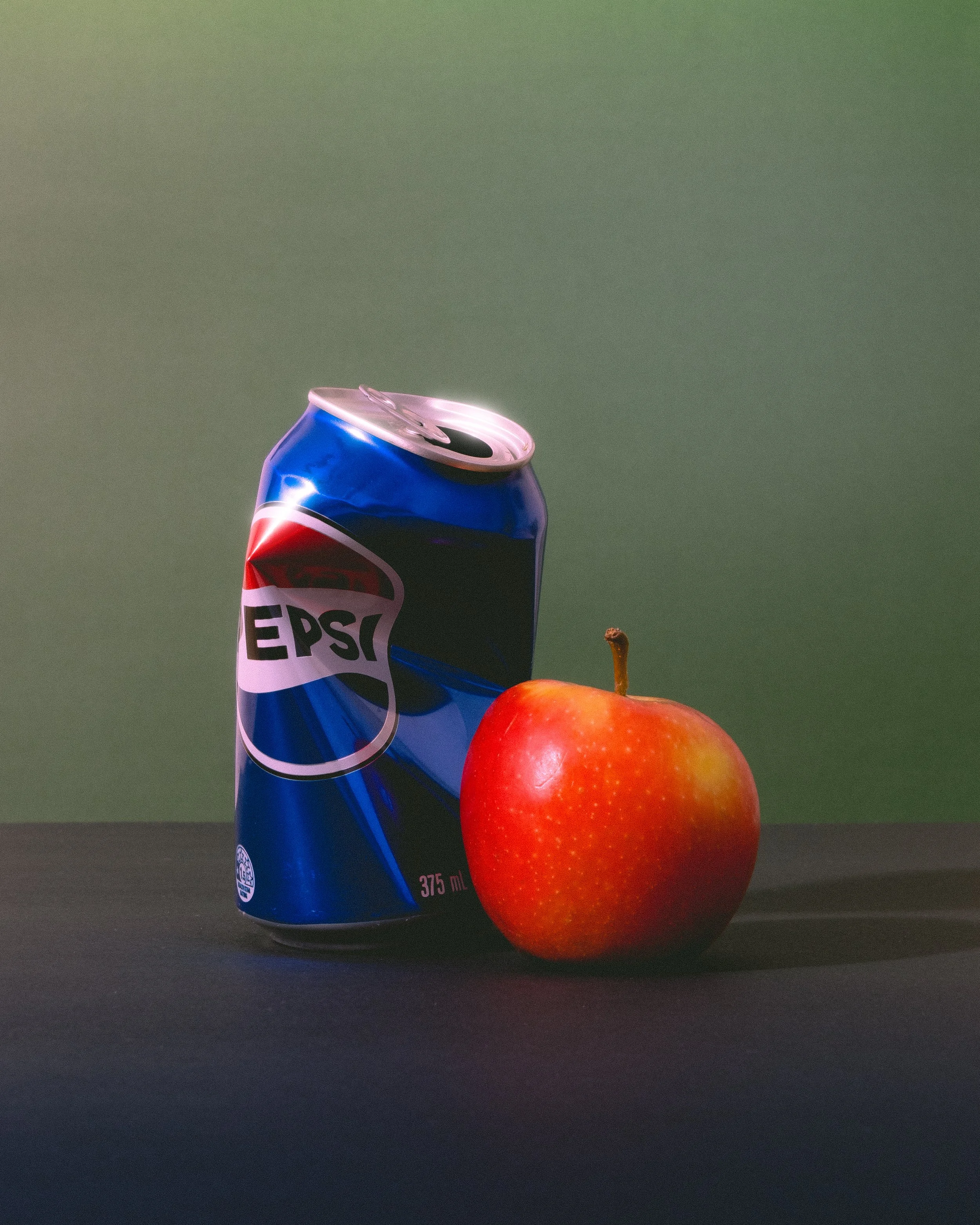 A can of Pepsi soda and a red apple placed on a dark surface with a green background.