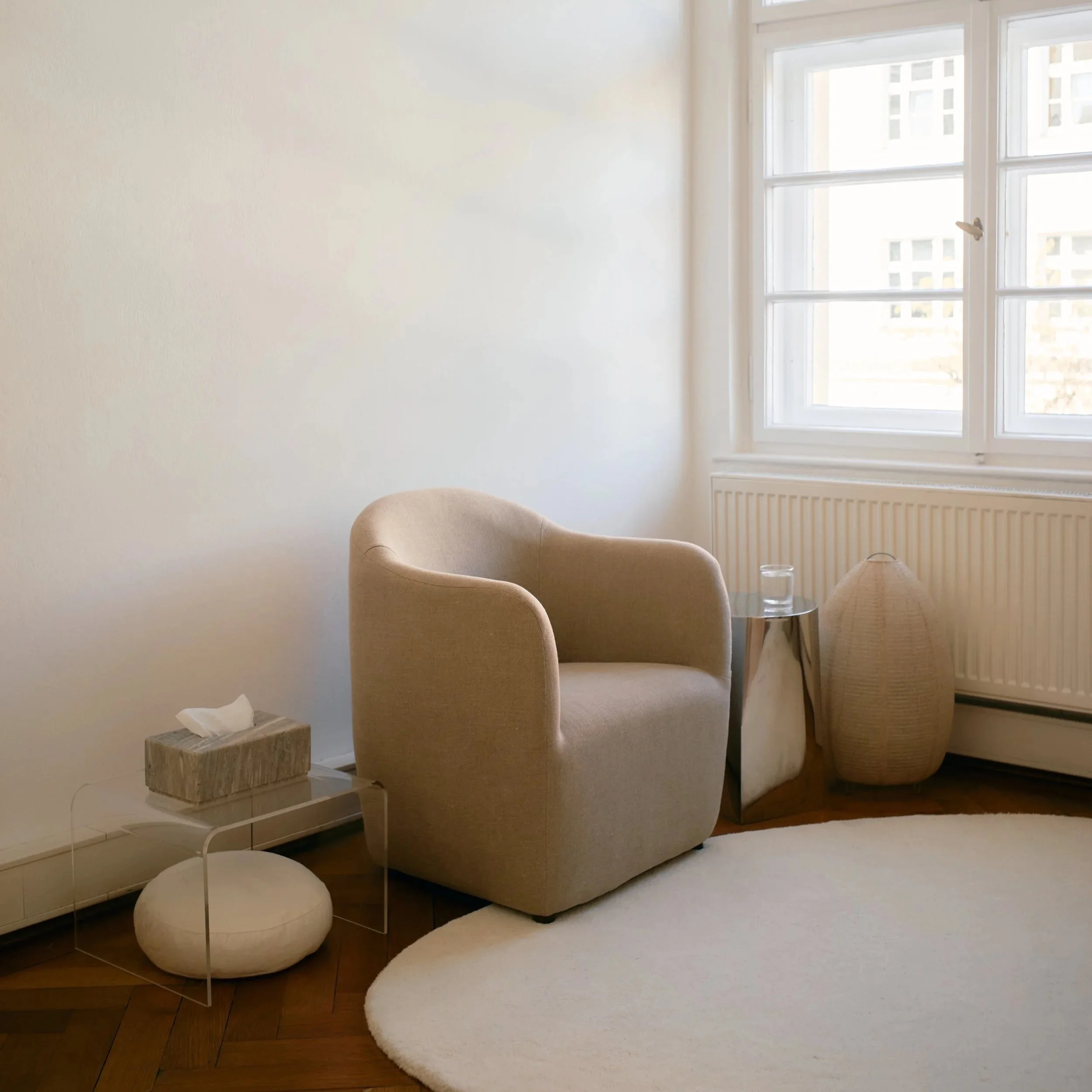 Helles, modernes Wohnzimmer mit beigefarbenem Sessel, Beistelltisch, Zimmerpflanze, großem Fenster und weißem Rundteppich.