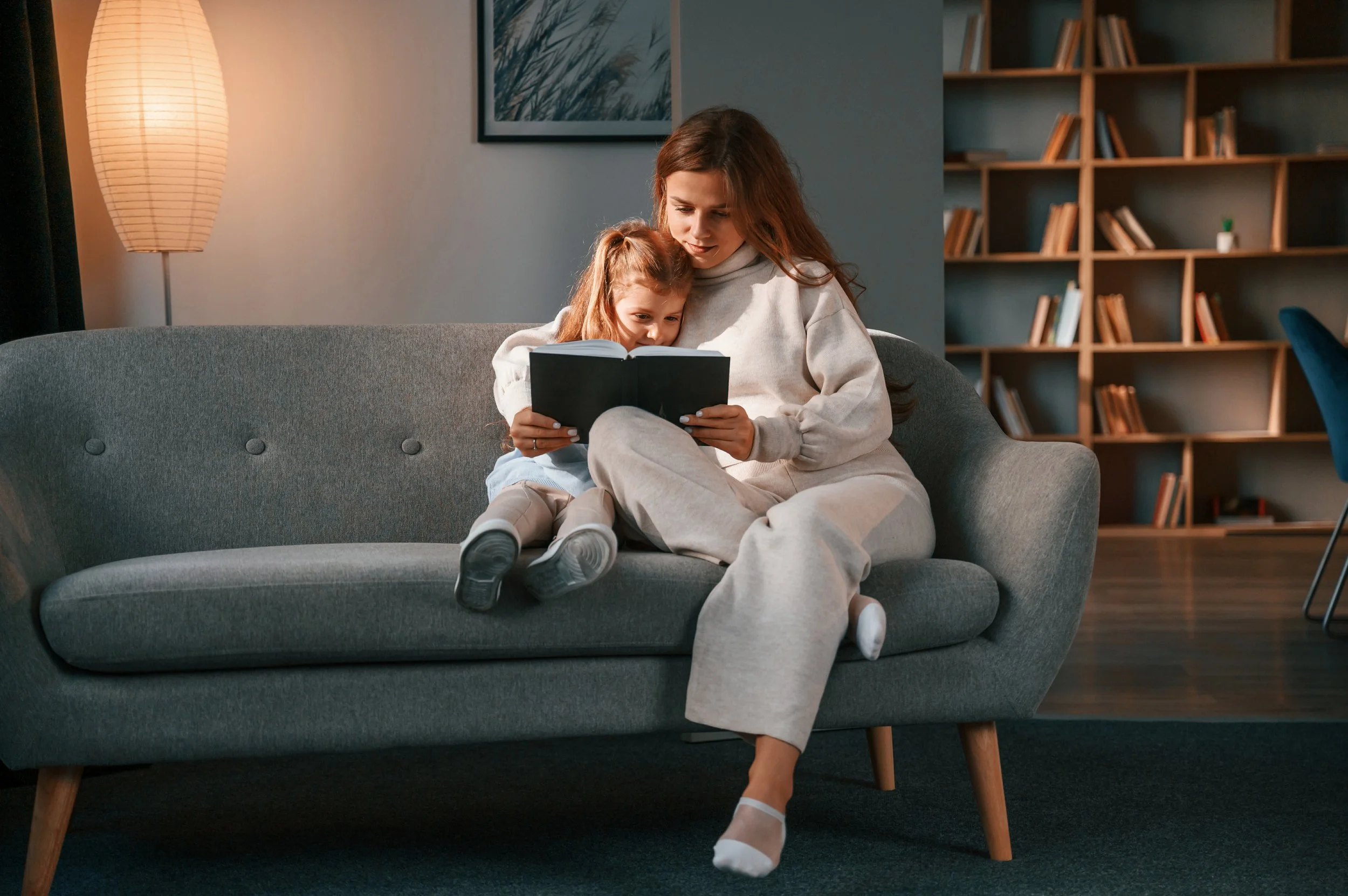 Woman and girl reading on a gray sofa in cozy living room and a bookshelf in the background.