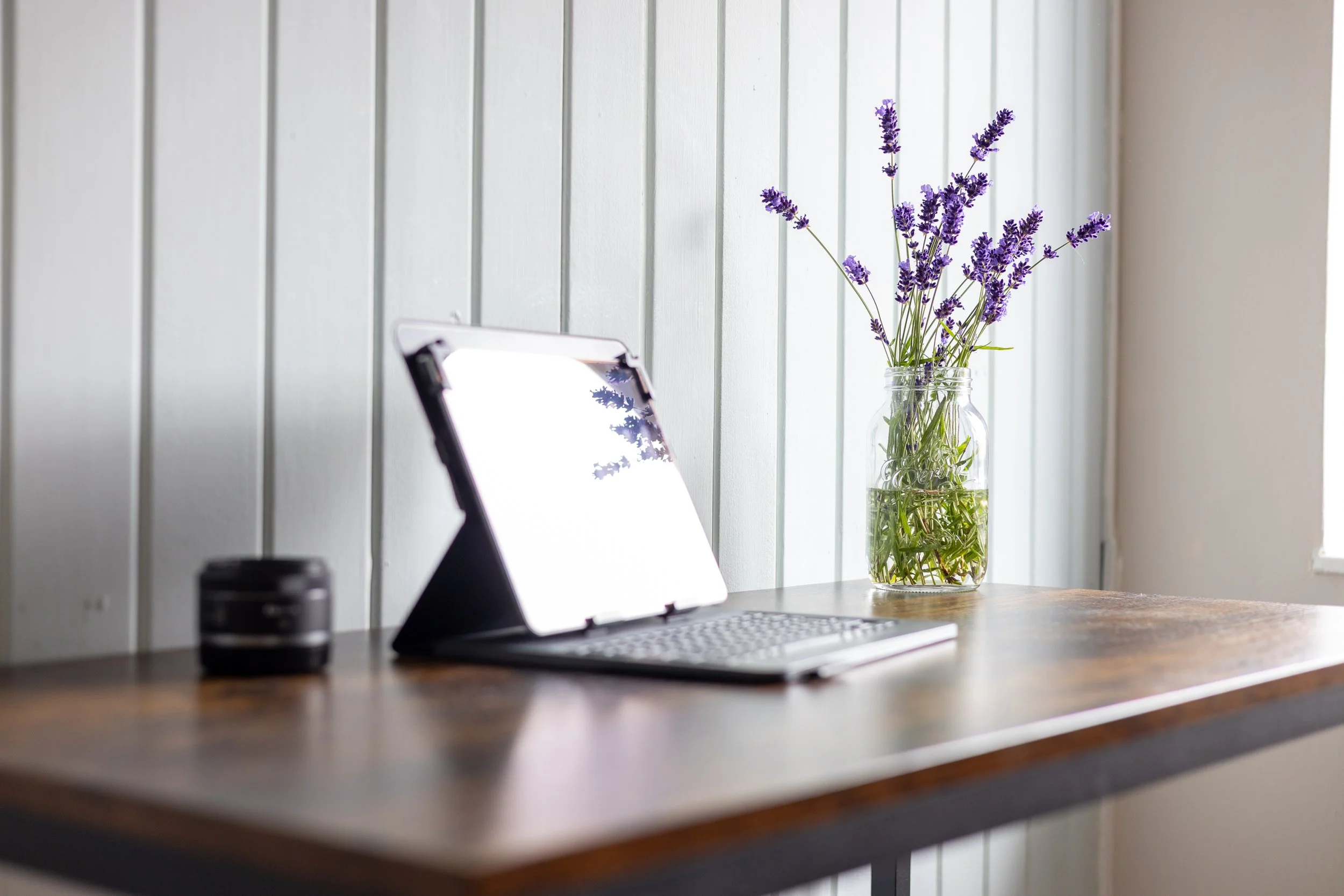 A wooden desk with a tablet, a camera lens, and a glass jar of purple lavender flowers.