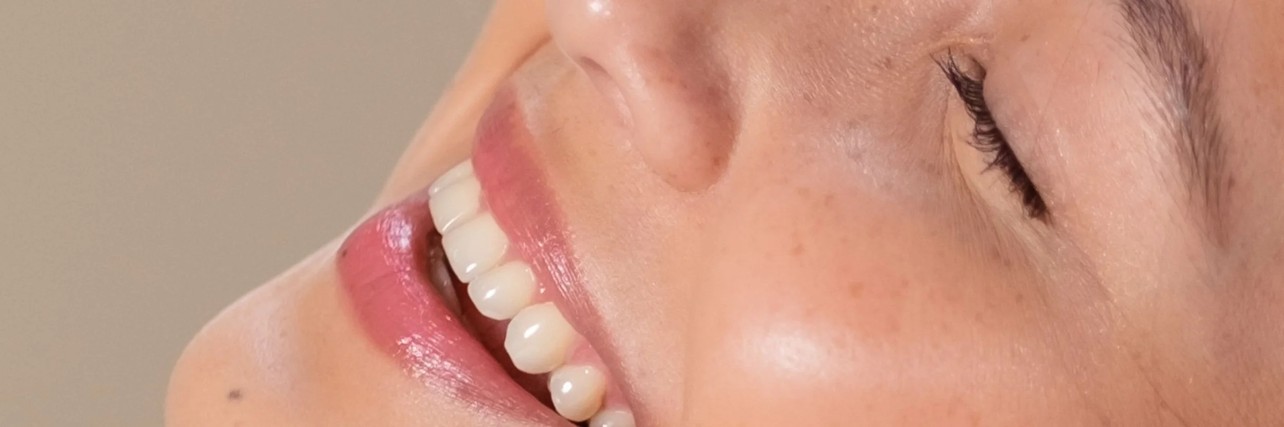 Close-up of a woman smiling, showing her white teeth and pink lips, with her eyes closed and skin glowing.