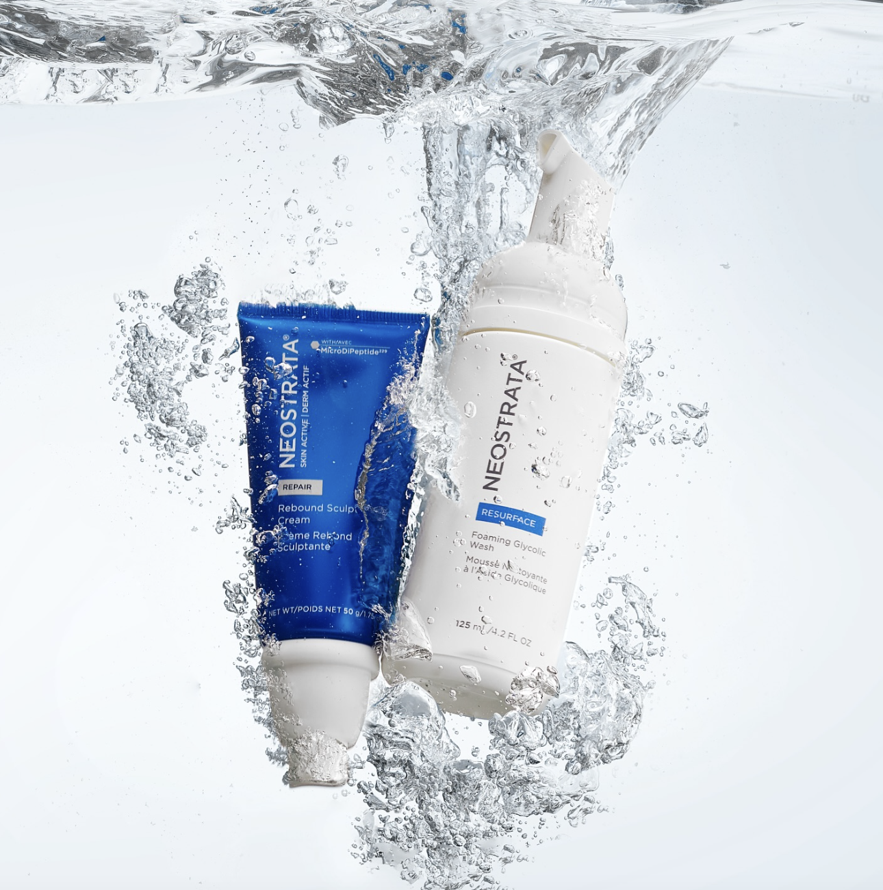 Two skincare products, a tube of NeoStrea Rebound Sculpt Cream and a bottle of NeoStrea Resurface Foaming Glycolic Wash, submerged in water with bubbles and splashes around them.