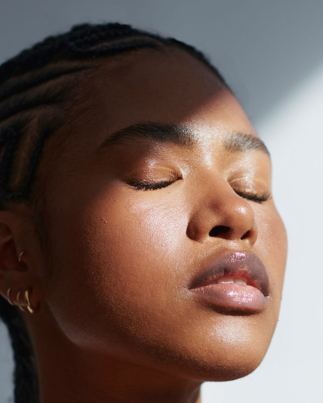 Close-up of a woman with dark skin and braided hair, with eyes closed and glossy lips, illuminated by natural light.