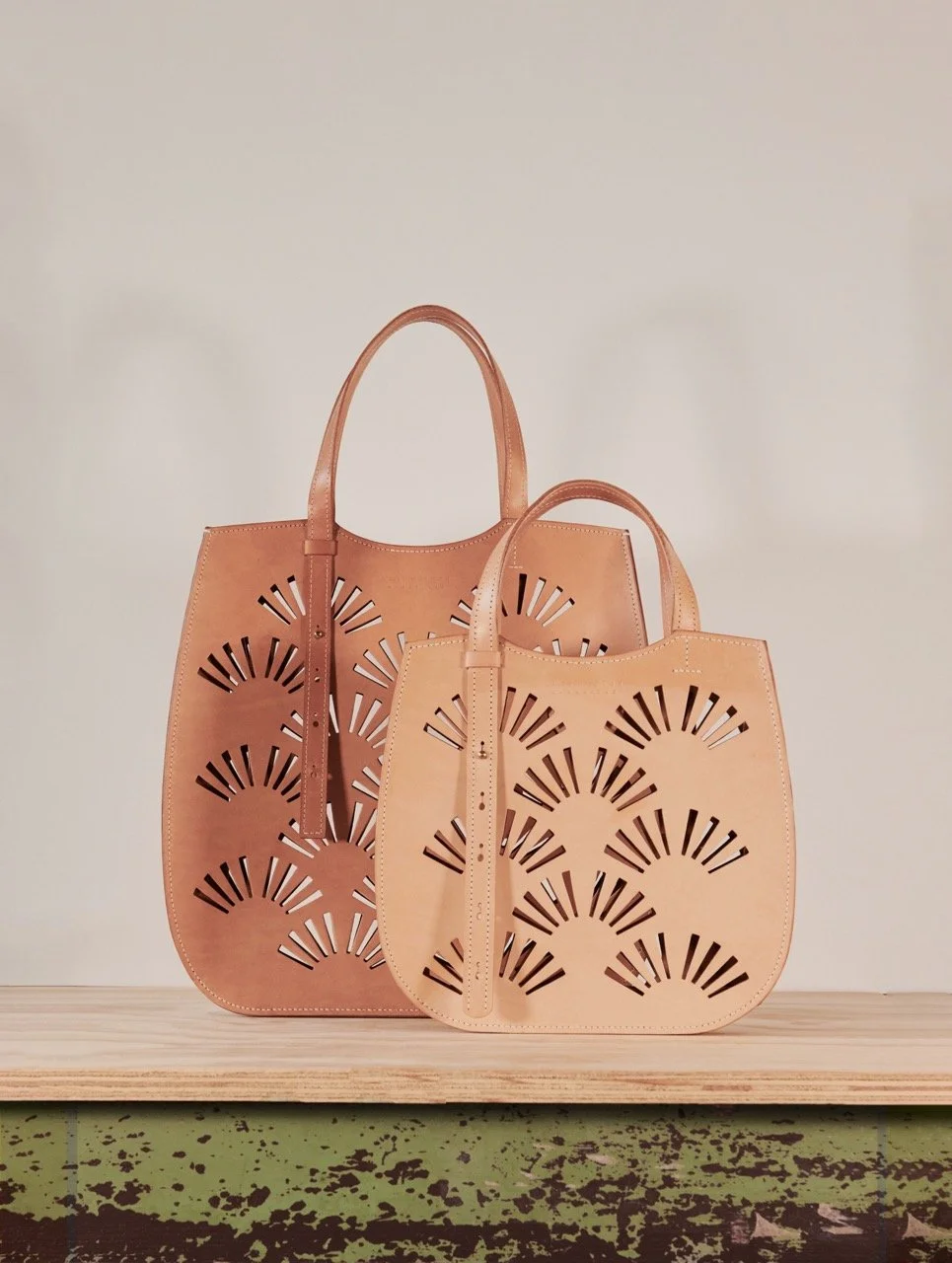 Two leather tote bags with cutout sunburst design, one in light beige and the other in darker brown, placed on a wooden surface against a plain wall.