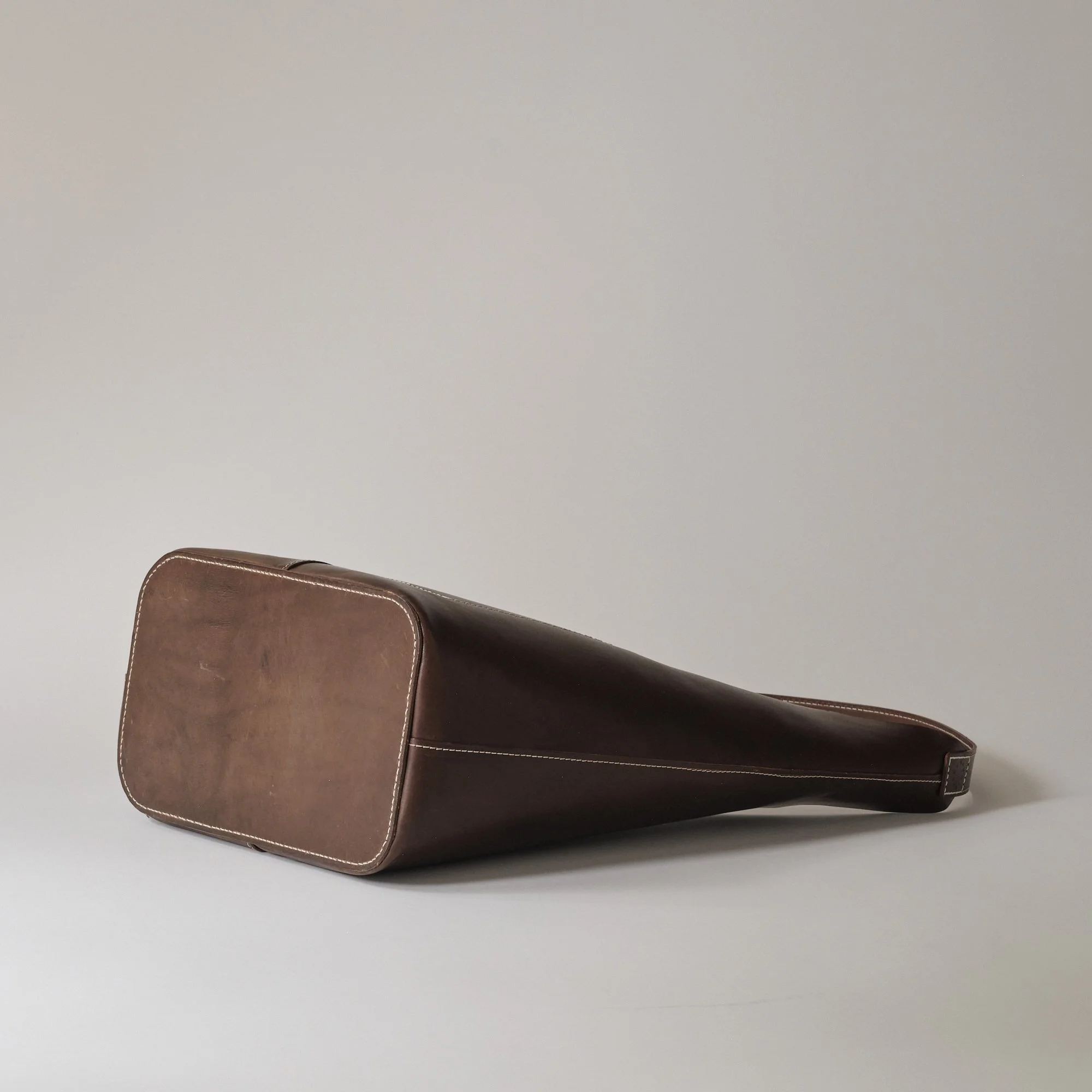 A brown leather bag with a rectangular bottom and a tapered body, lying on a white surface against a light gray background.