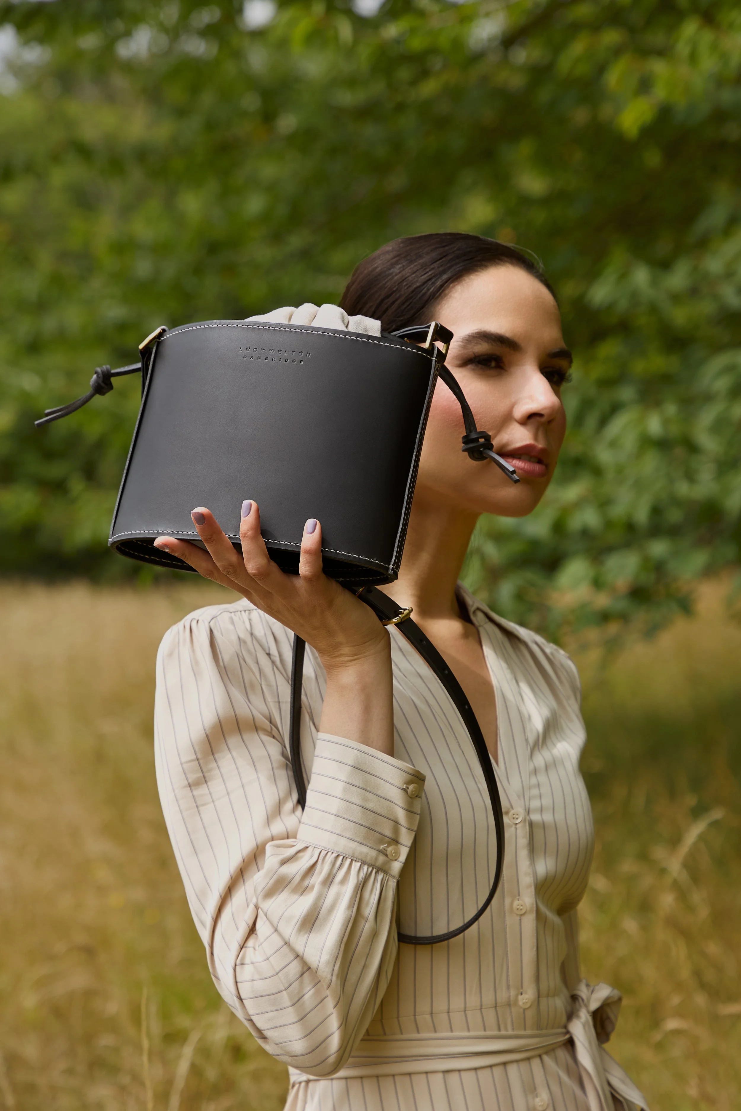 A woman outdoors holding a black handbag over her shoulder, surrounded by green trees and grass.