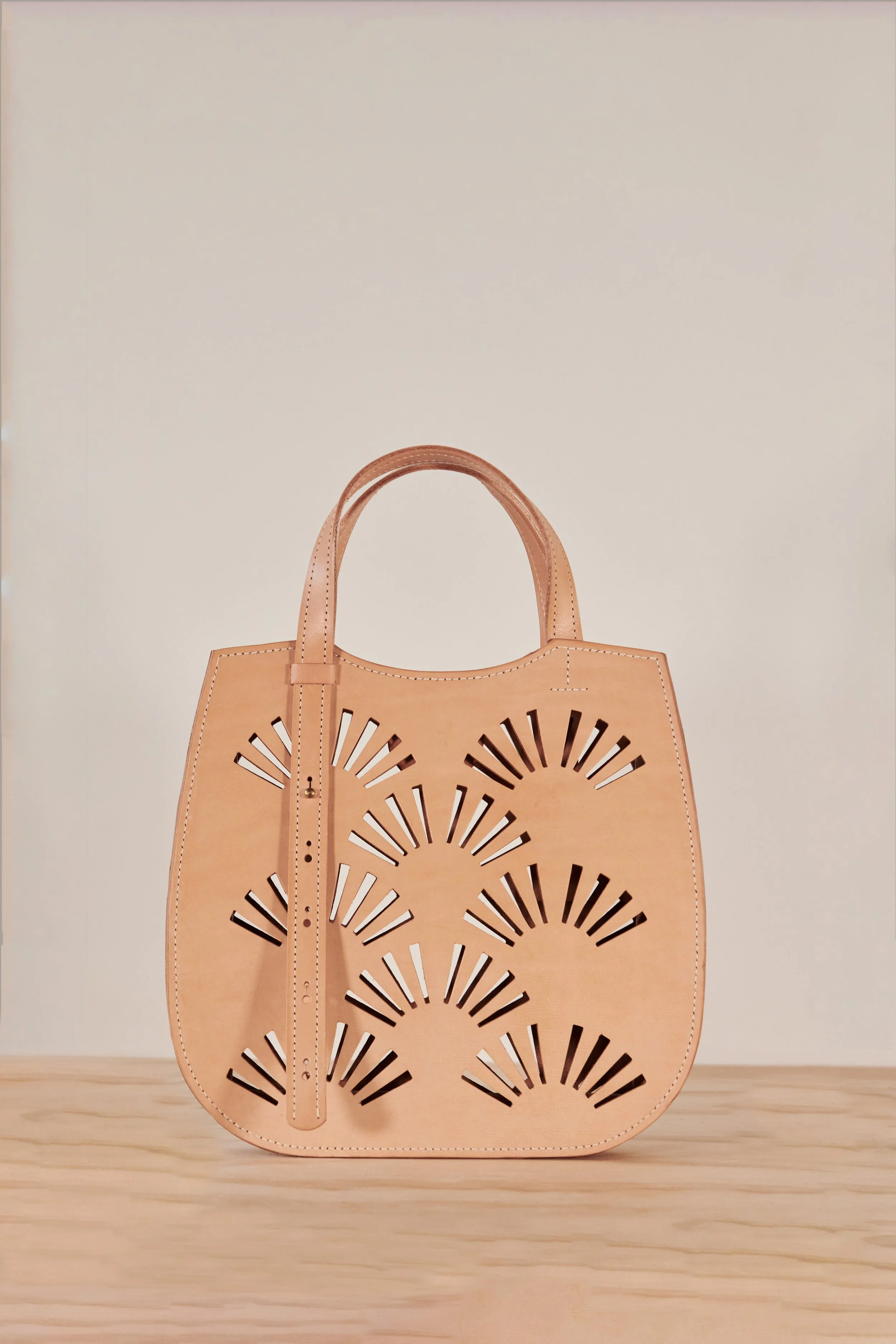 A tan leather handbag with multiple cutout fan-shaped patterns, resting on a wooden surface with a plain off-white background.
