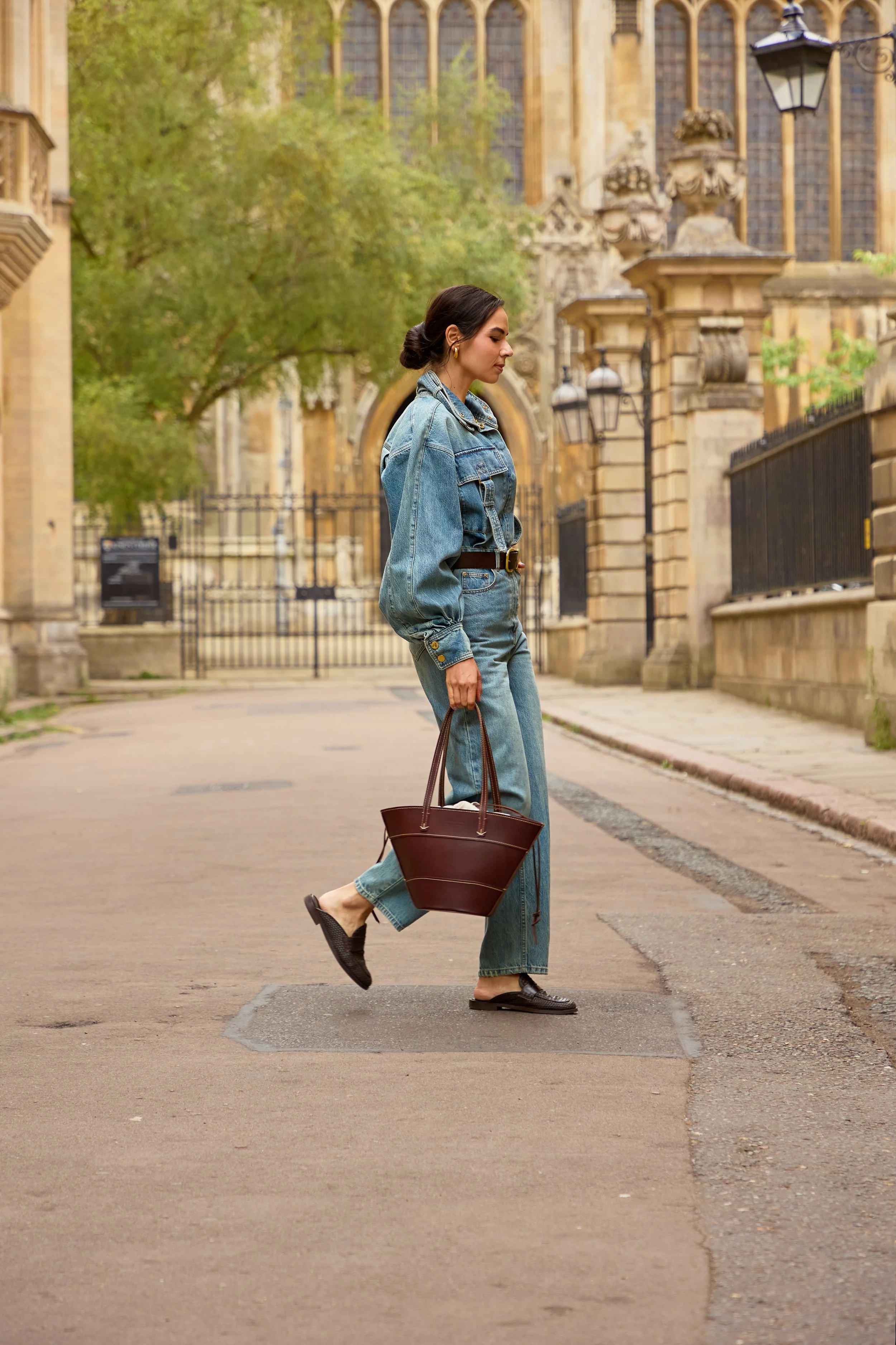 A woman walking on a sidewalk near a historic building, wearing a denim jacket and jeans, carrying a large brown handbag, with an overcast sky and green trees in the background.