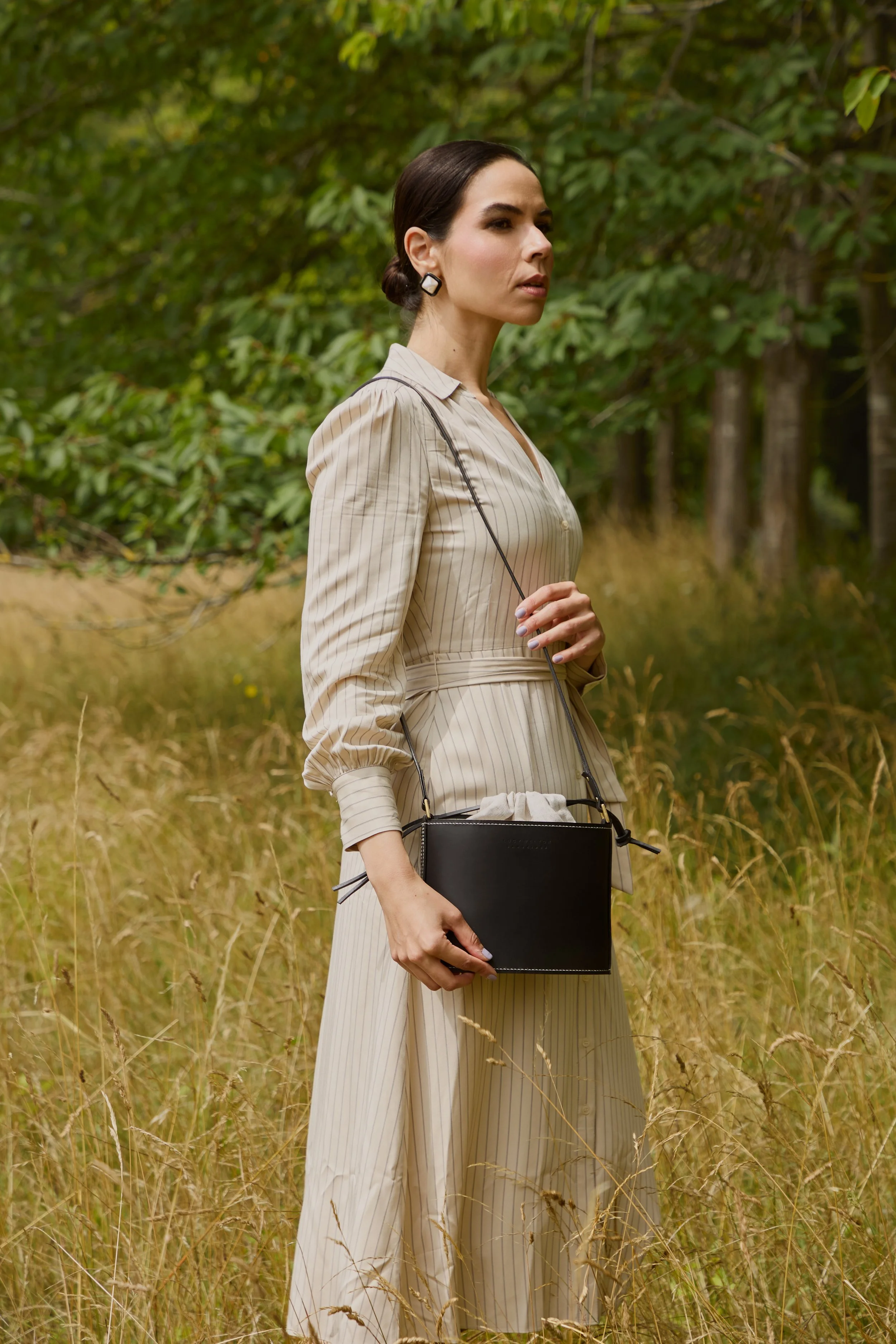 A woman standing in a field with tall grass and trees in the background, wearing a beige striped outfit and holding a black purse.