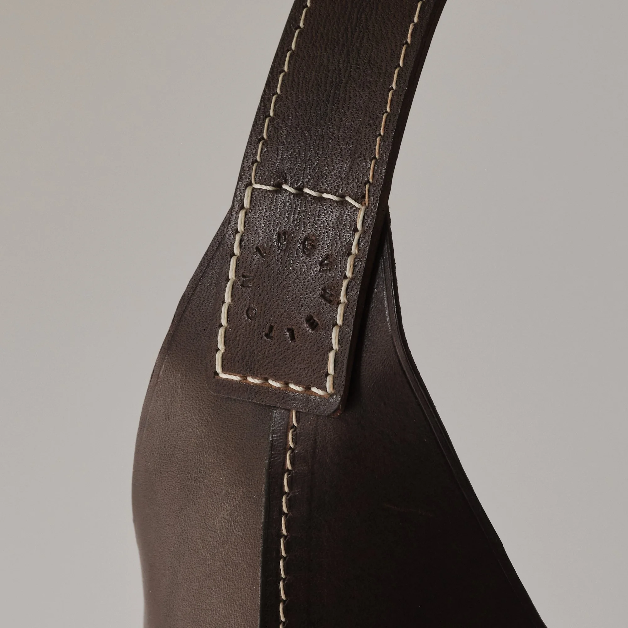 Close-up of a dark brown leather strap with beige stitching and embossed brand lettering, attached to a piece of dark brown leather.