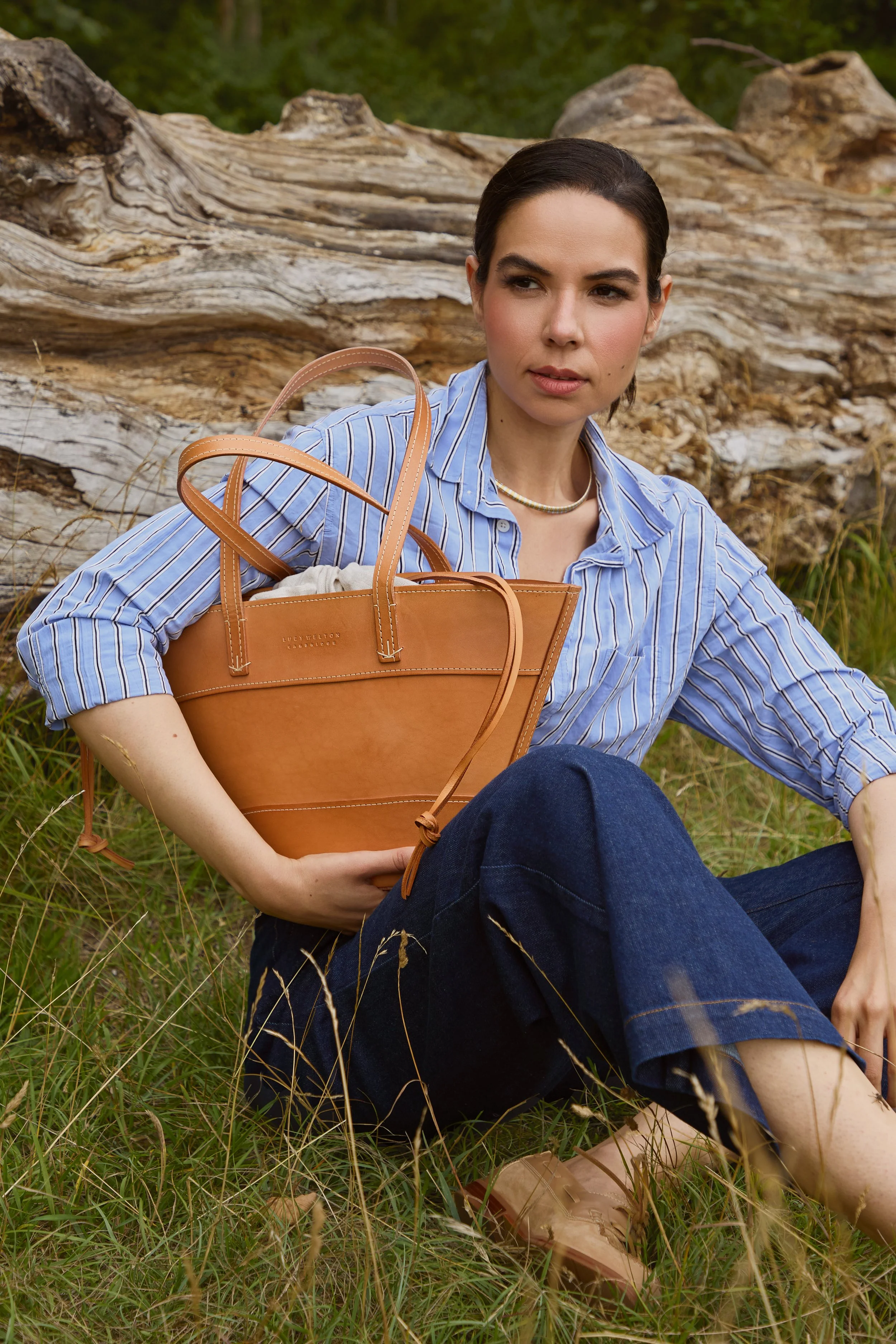 Woman sitting outdoors on grass near a fallen log, wearing a blue and white striped shirt, dark jeans, holding a tan leather tote bag.