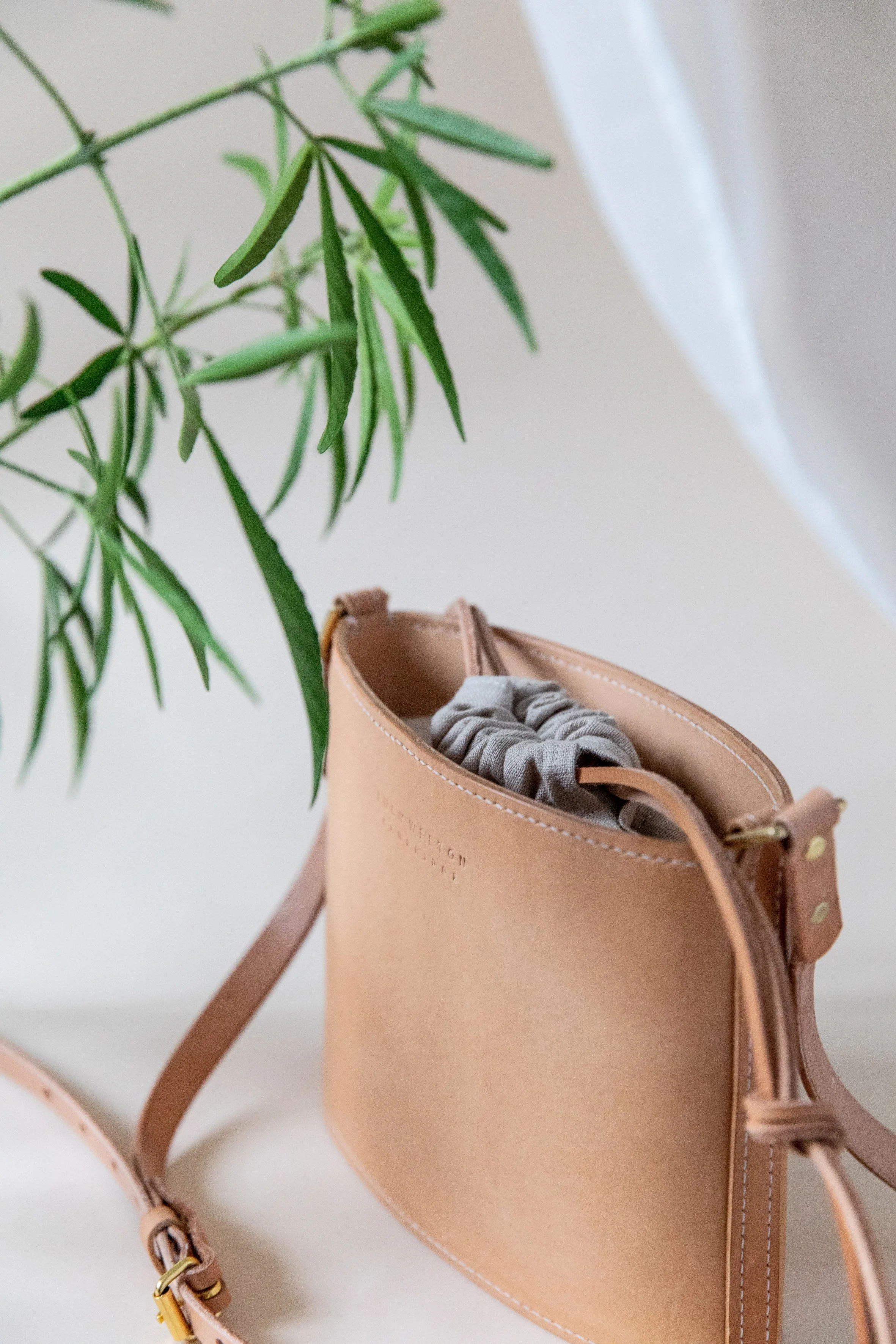 A tan leather handbag with a grey scrunchie inside, placed below a green leafy plant in a well-lit minimalistic setting.