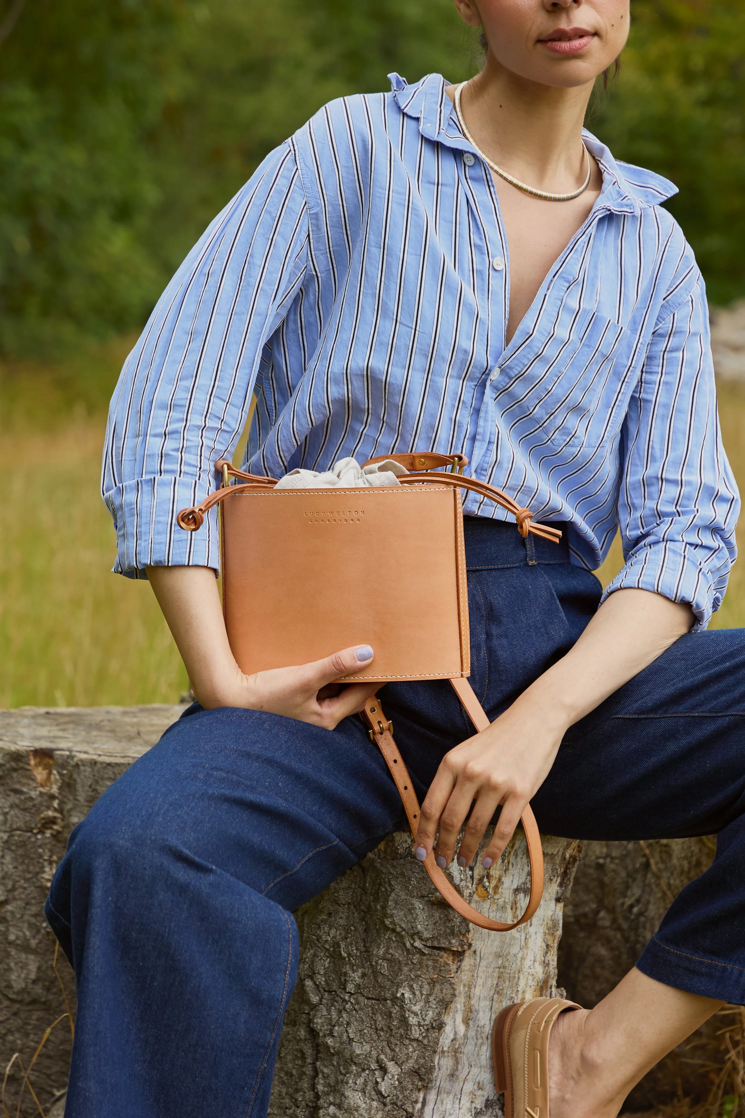 A woman sitting outdoors on a tree stump, holding a tan leather handbag, wearing a blue striped shirt and jeans.