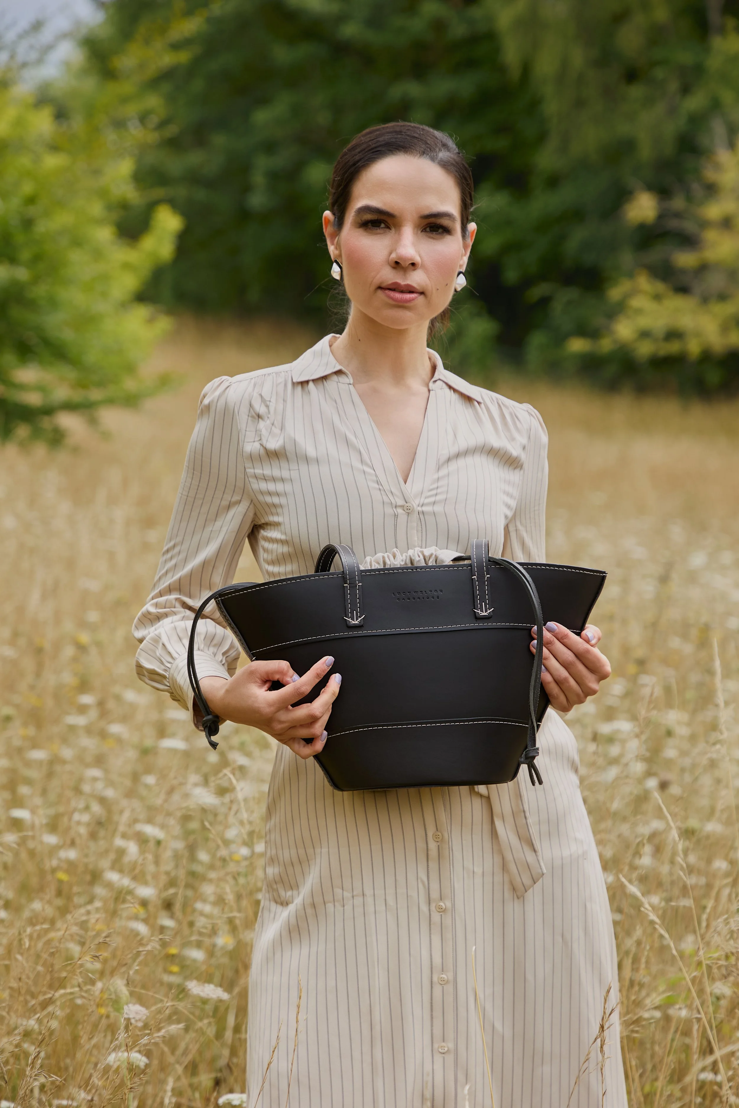 A woman standing in a field of tall grass and trees, holding a large black designer handbag, wearing a beige striped dress with puffed shoulders, earrings, and light makeup.