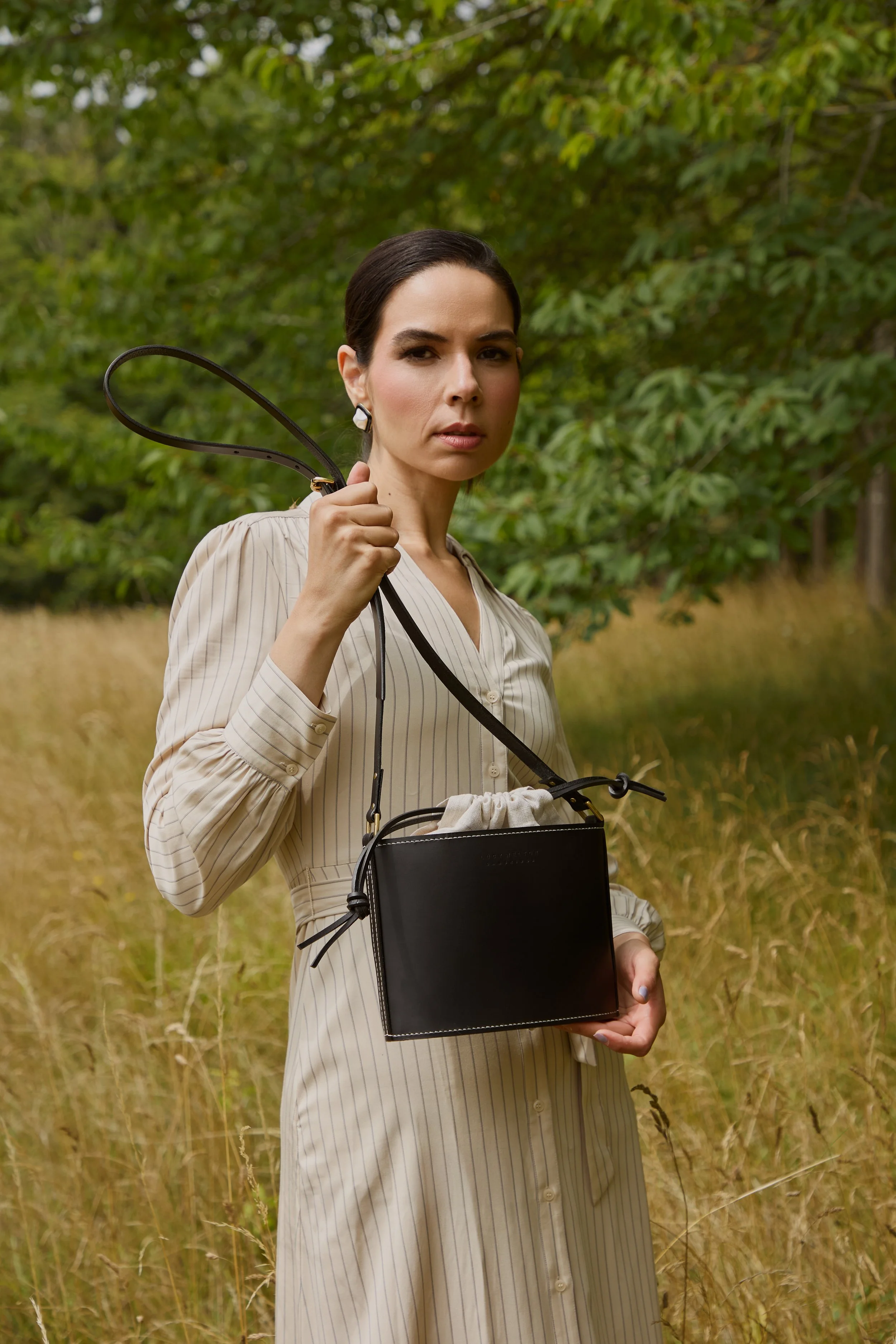 A woman in a light-colored, striped dress stands outdoors in a grassy area with trees in the background, holding a black handbag with a strap over her shoulder.