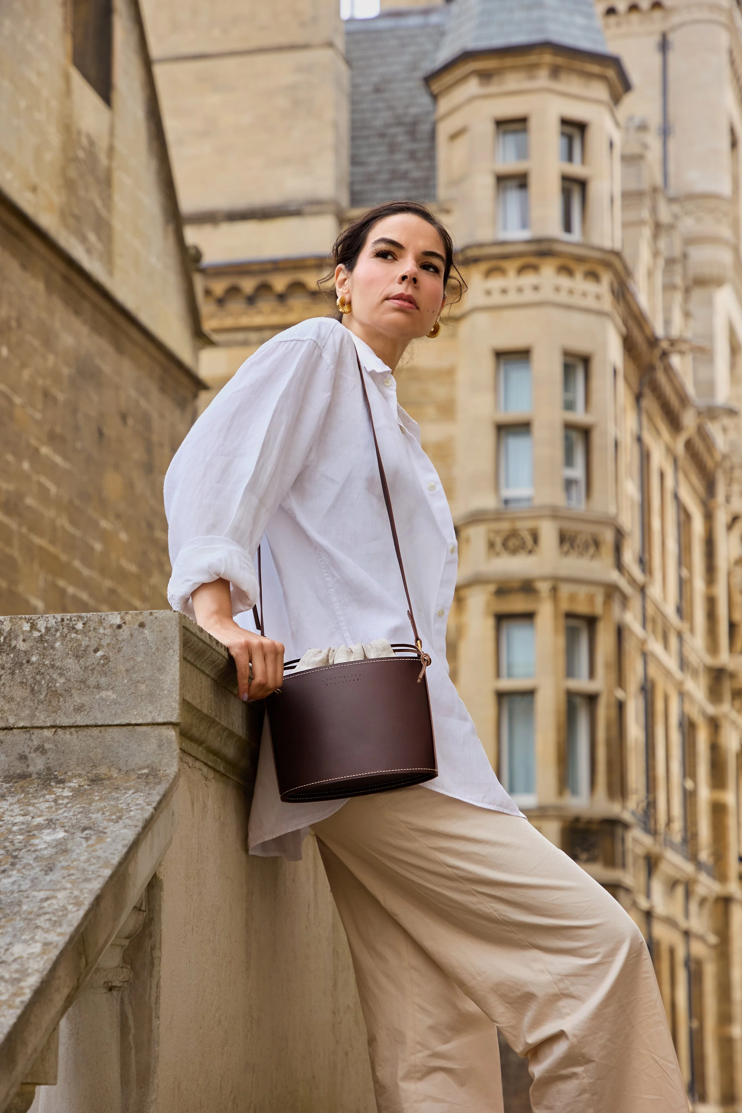 A woman wearing a white shirt and beige wide-leg pants leaning against a stone ledge in front of a historic building, holding a dark brown handbag with a strap over her shoulder, with an urban cityscape in the background.
