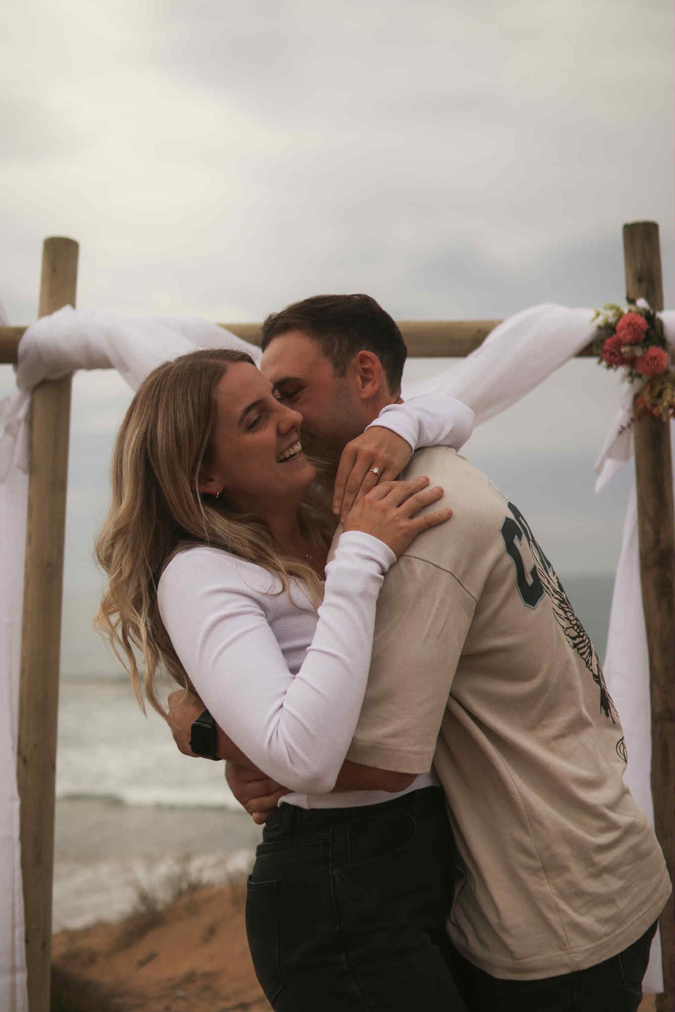 A couple embraces on the beach in front of a wooden arch decorated with pink flowers, sharing a kiss.