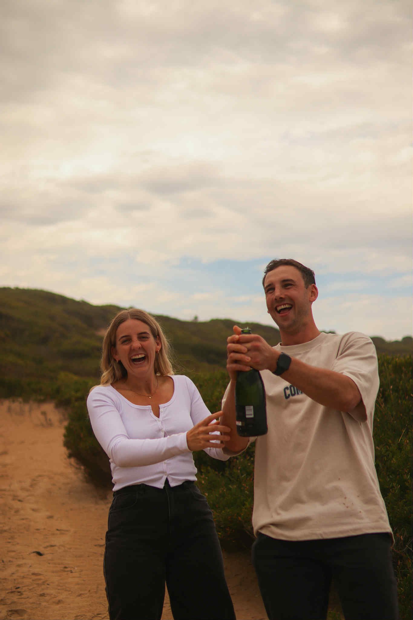 A man and woman smiling outdoors on a cloudy day, with the man opening a bottle of champagne or sparkling wine.