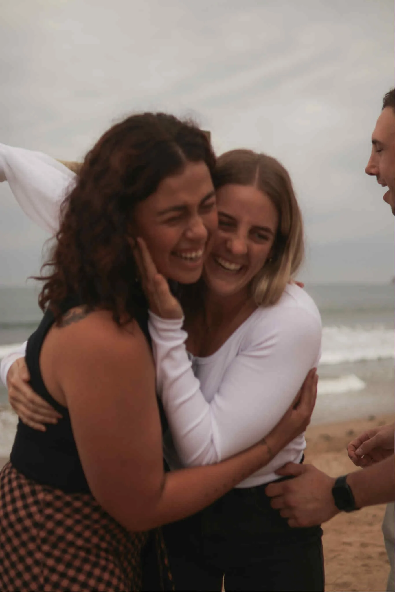 Three women and one man sharing a hug and smiling on a beach during cloudy weather.