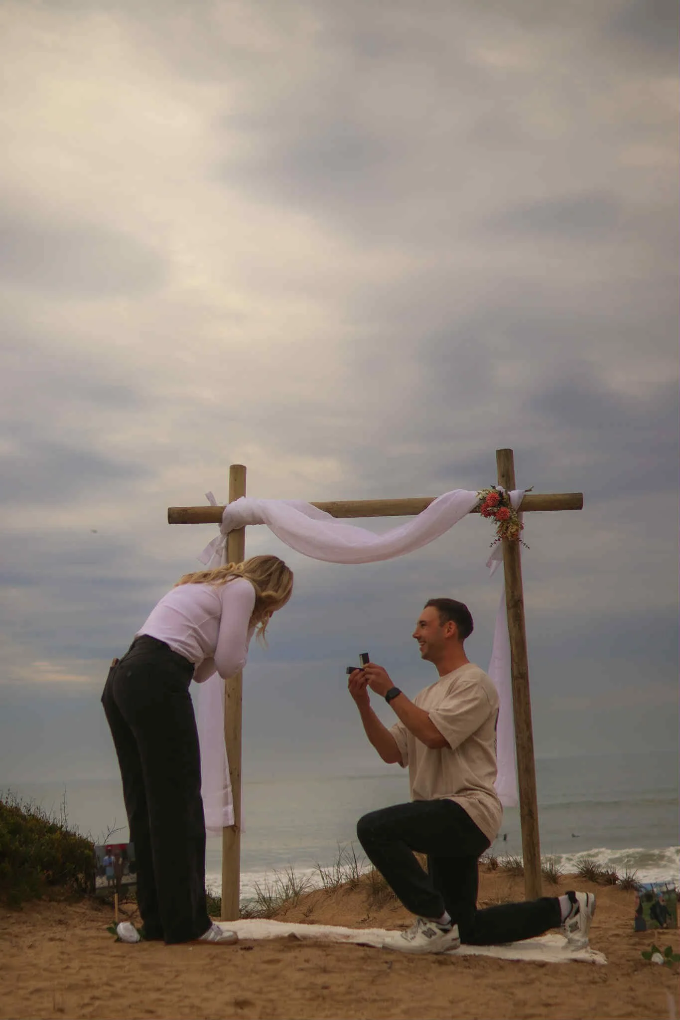 A man is kneeling on one knee holding a ring box, proposing to a woman who is bent forward, smiling, during an outdoor wedding ceremony. They are under a wooden arch decorated with flowers and fabric. The scene is set on a beach with dunes and a cloudy sky.