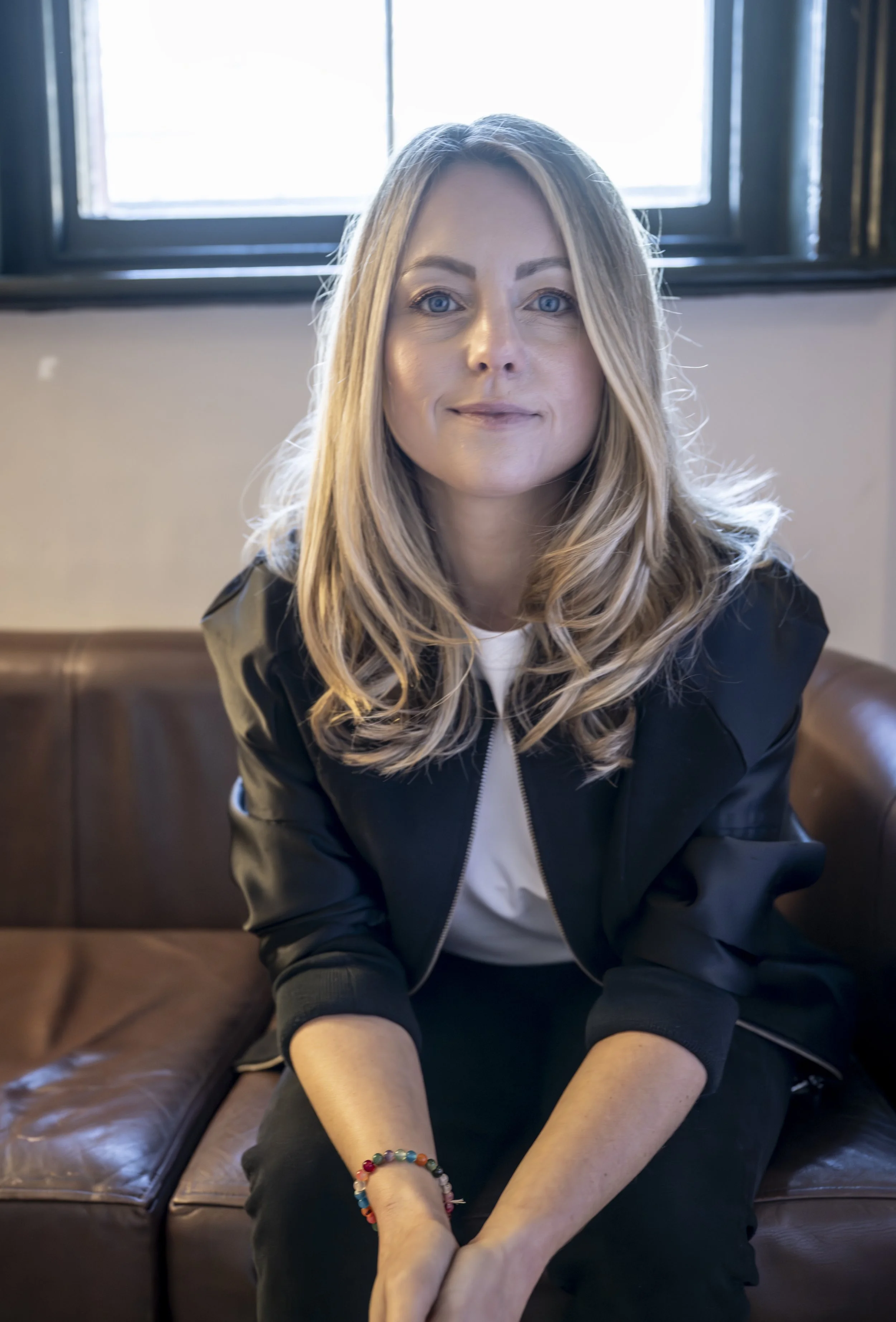 Coach, consultant & strategist for the music, tech and creative industries, Nicola Fuller, wearing a black jacket, sitting on a leather couch.
