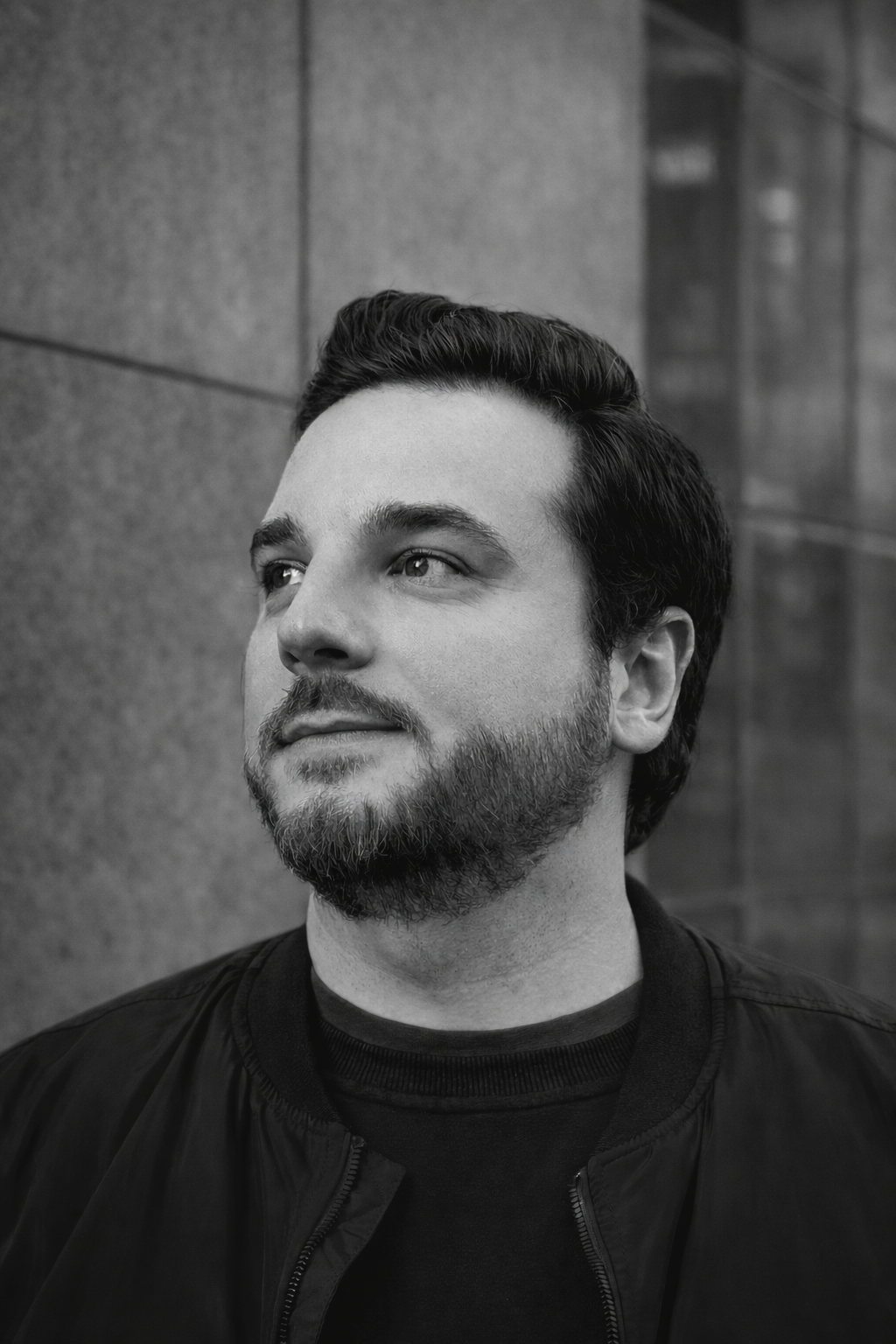 Black and white portrait of a man with styled hair and a beard, looking to the side, standing outdoors near a stone wall.