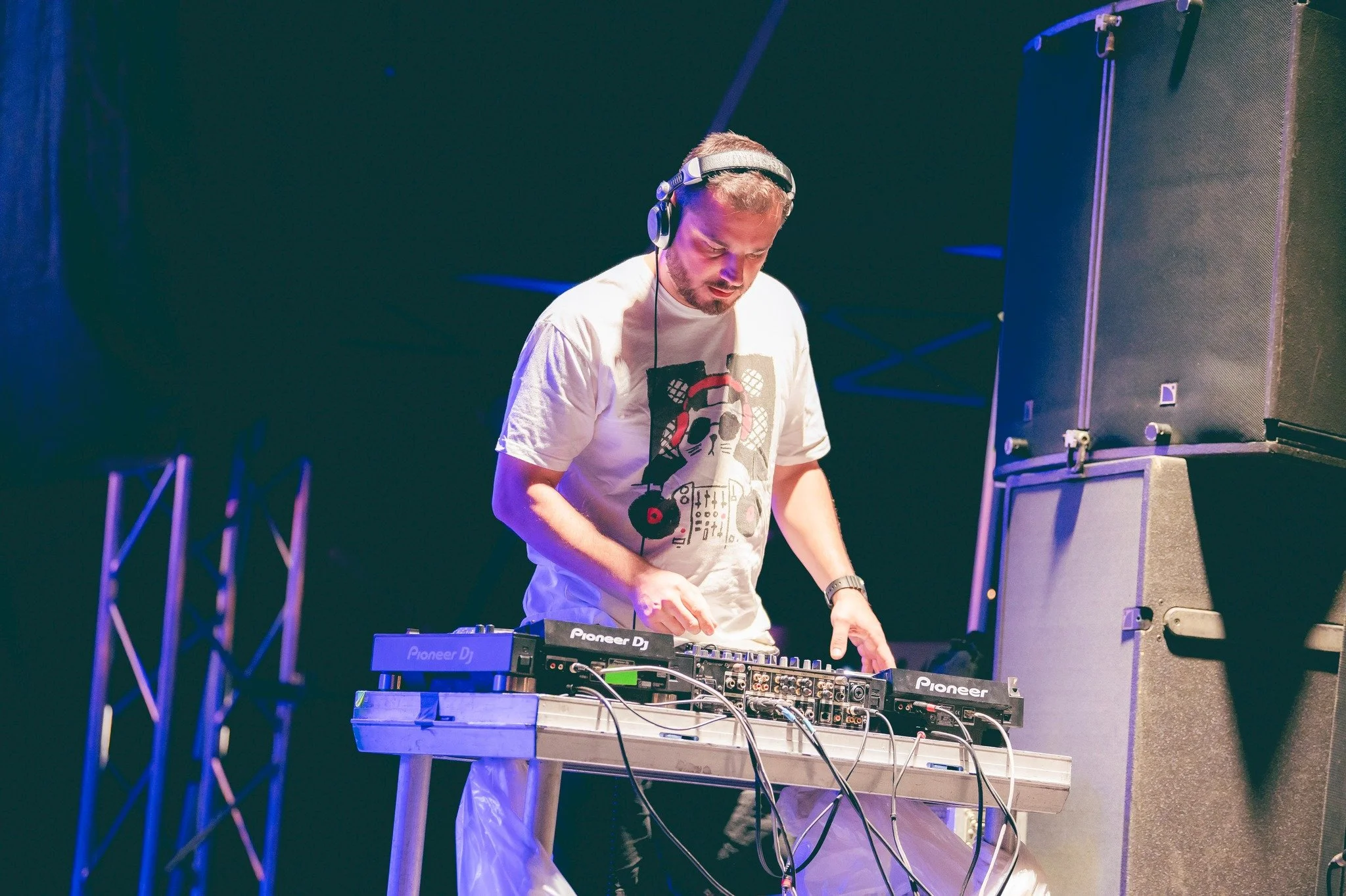 A DJ wearing headphones, a white T-shirt with a graphic of a skull and turntables, plays music on a Pioneer DJ controller on stage at a nightclub or music event.