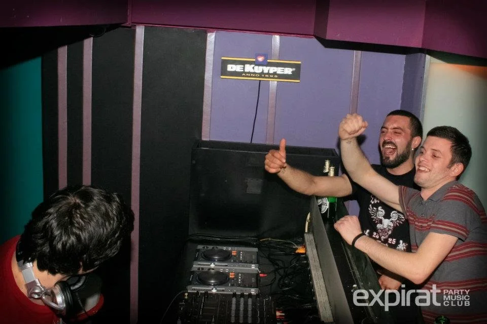 Two men smiling and celebrating with fists raised in front of DJ equipment at a party or club. A person with headphones is looking at them. The background has colorful wall panels and a sign that reads 'DE KUYPER ANNO 1880'.