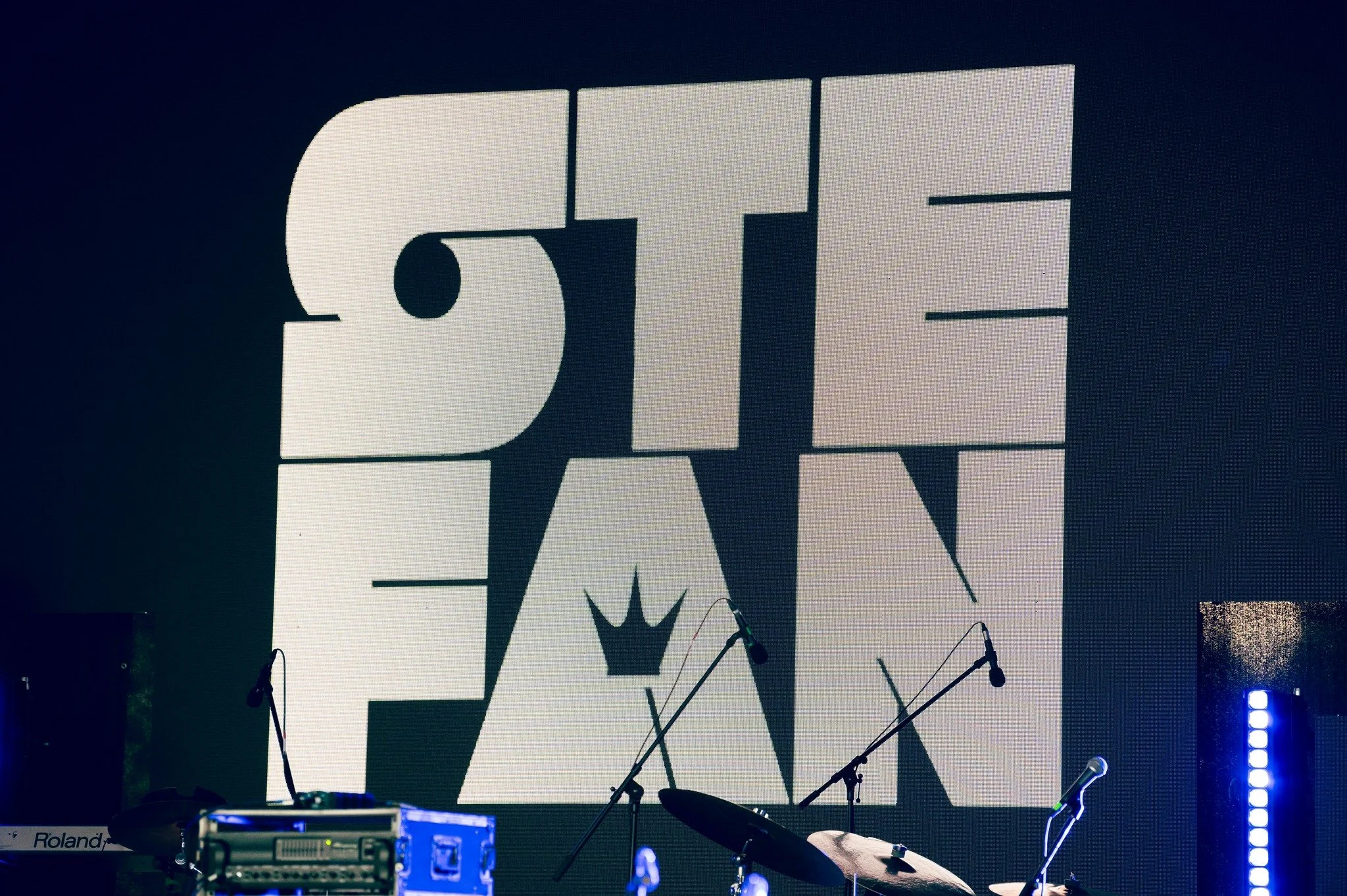 Stage setup with musical instruments, microphones, and a large screen displaying the text 'THE' with a crown symbol in the middle in black and white.