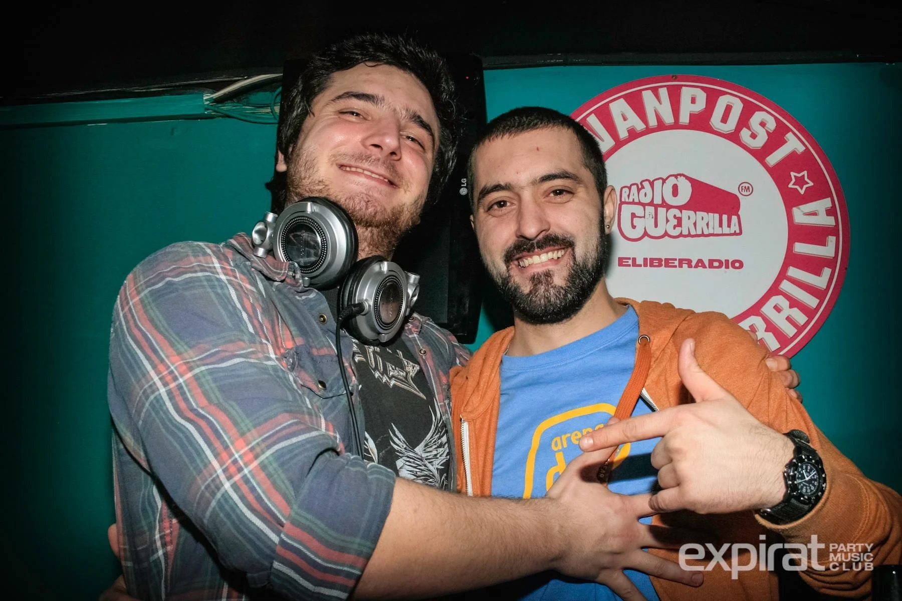 Two men smiling and embracing each other at a party, one wearing headphones around his neck and the other pointing at him. A banner with logo 'Radio Guerrilla' and text 'Eliberadio' is in the background.