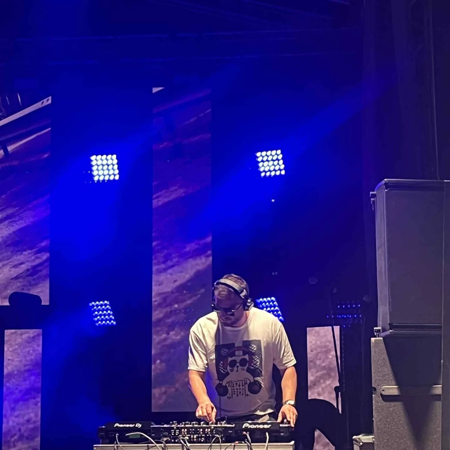 A DJ wearing a beige T-shirt with a skull graphic, headphones, and glasses, is performing on stage with a Pioneer DJ controller. The background features large digital screens displaying abstract patterns, illuminated by blue stage lighting.