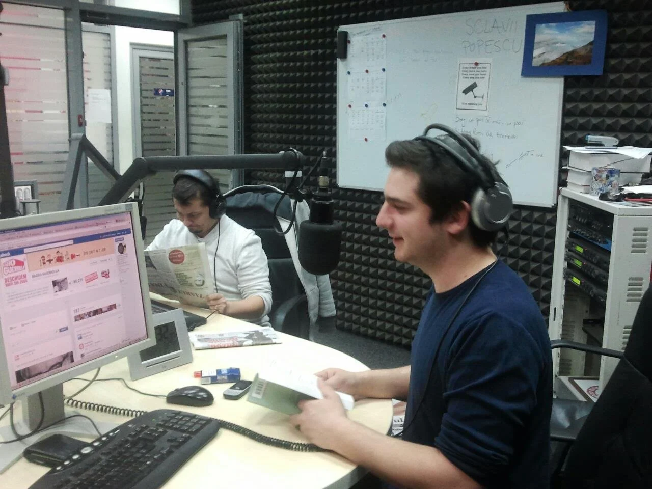 Two men wearing headphones in a radio studio, one reading a newspaper and the other holding a book, with a computer monitor, microphone, and sound equipment visible.