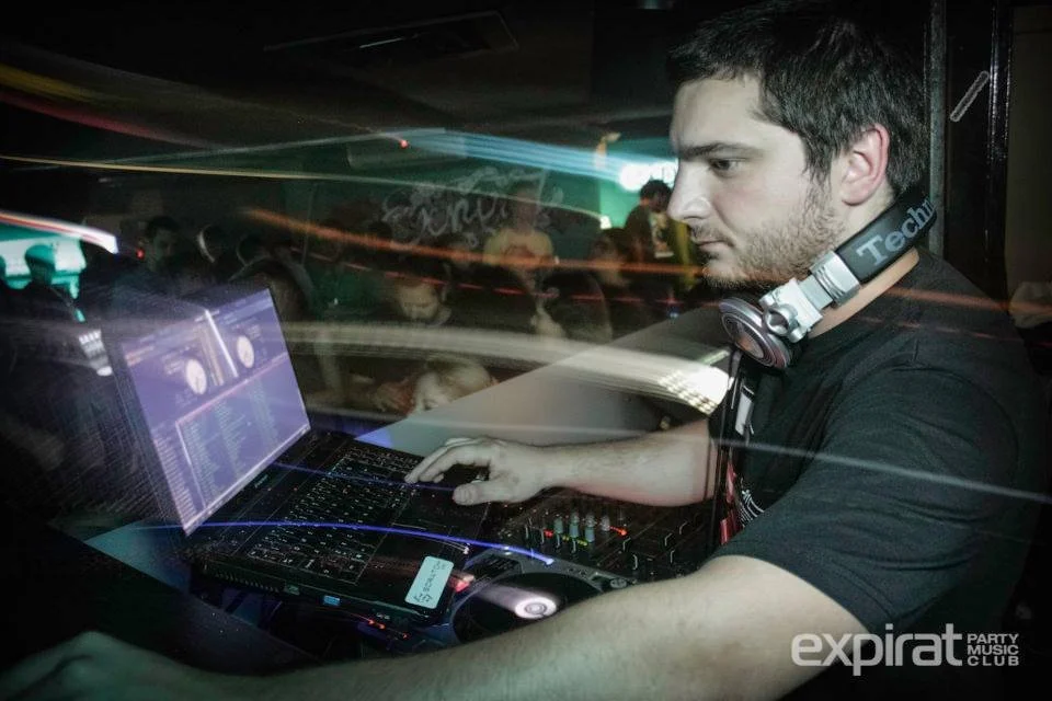 A DJ is mixing music on a laptop and DJ equipment at a party or nightclub, with people dancing in the background.