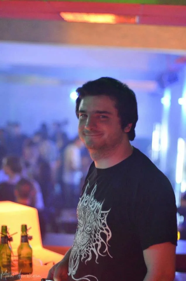 A young man with dark hair smiling at a nightclub or party with colorful neon lighting in the background. He is wearing a black T-shirt with a graphic design.