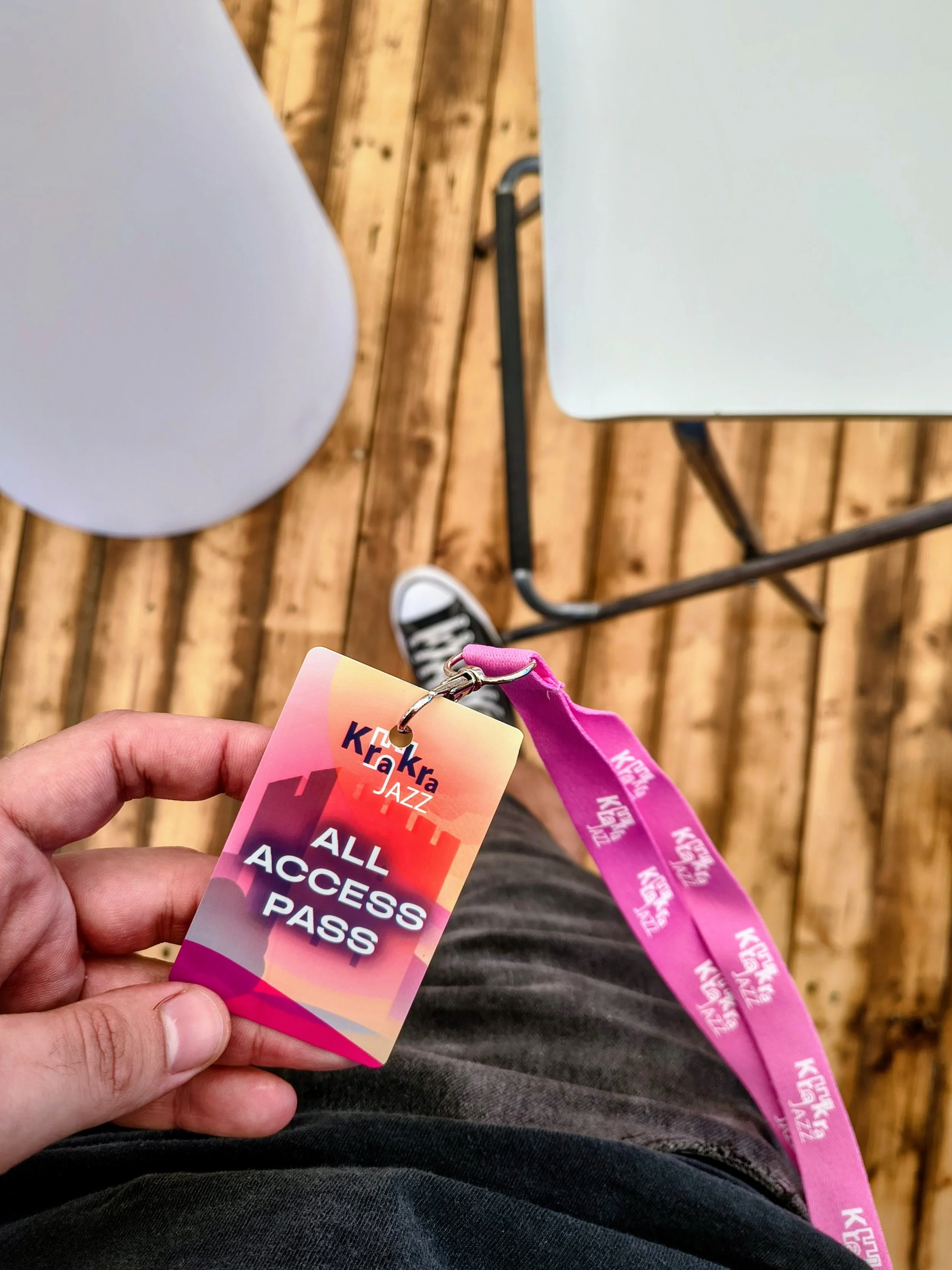 Person holding a colorful all-access pass badge for a jazz event, with a pink lanyard, sitting in a venue with wooden flooring, a blue chair, and a white table nearby.