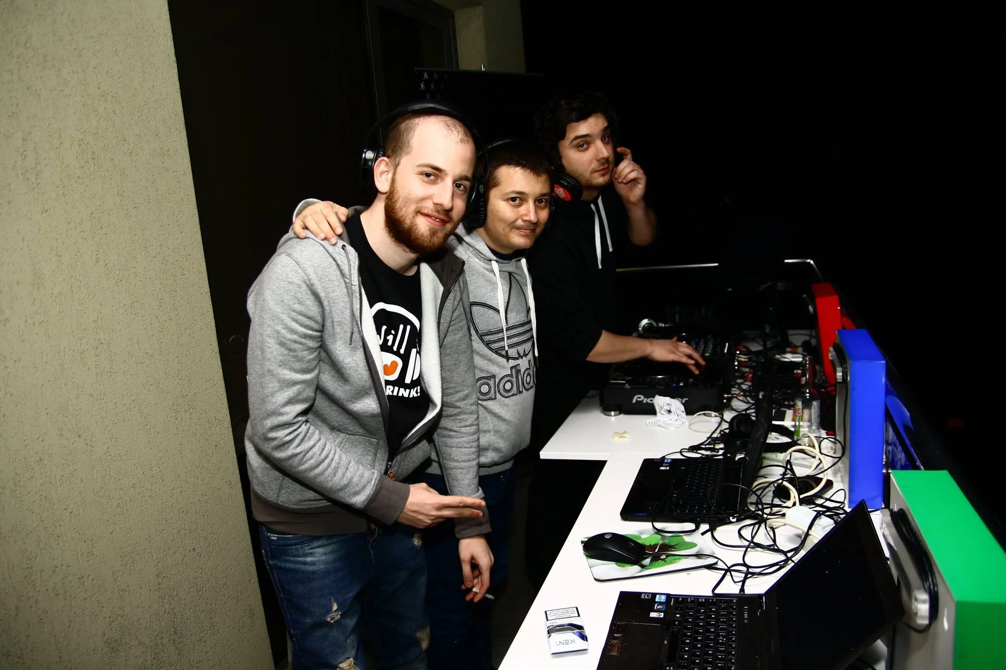Three young men wearing headphones stand together at a DJ setup with laptops, mixing equipment, and monitors in a dark room.