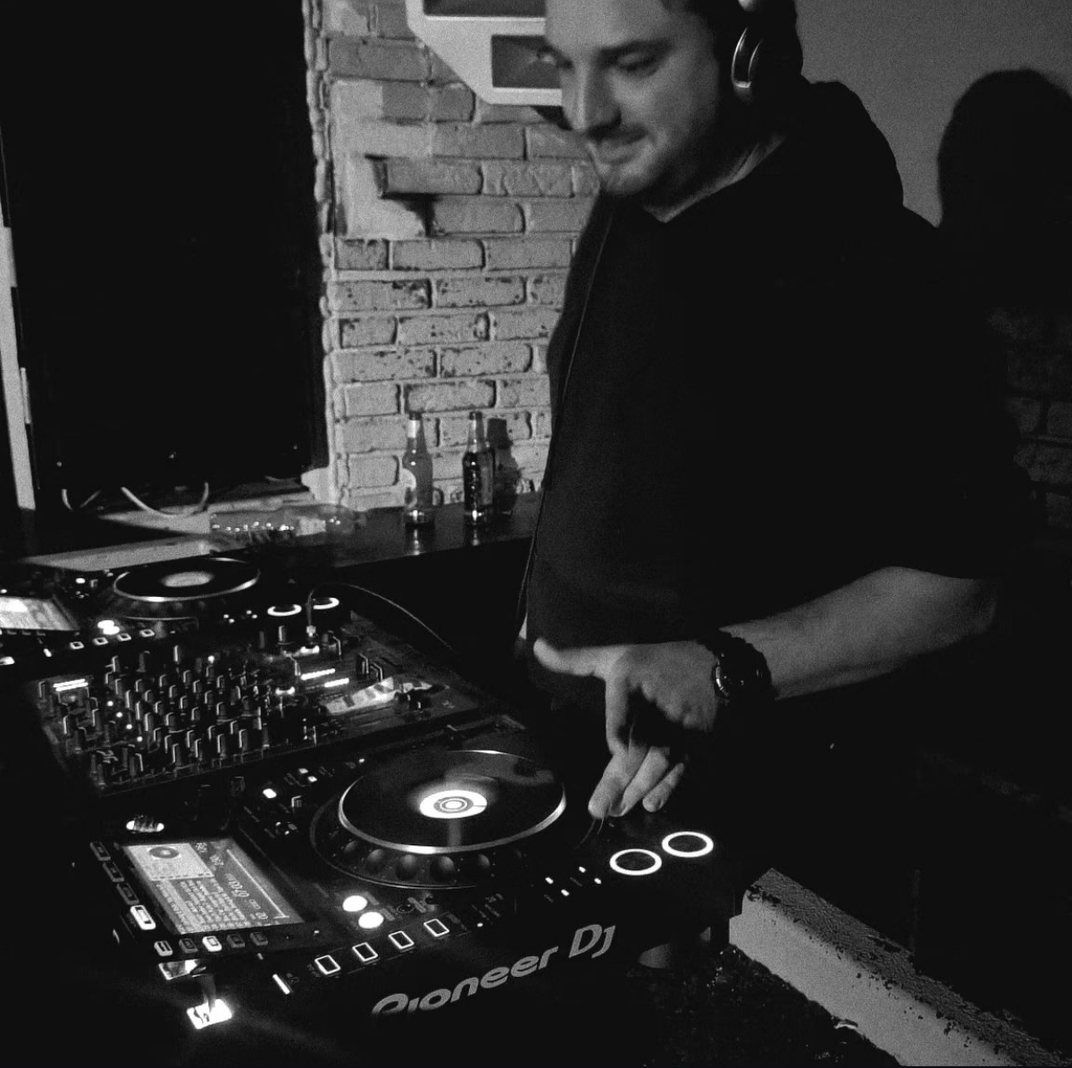 A person wearing headphones is DJing with a Pioneer DJ mixer, set up on a table in front of a brick wall, with a few bottles on a shelf behind them.