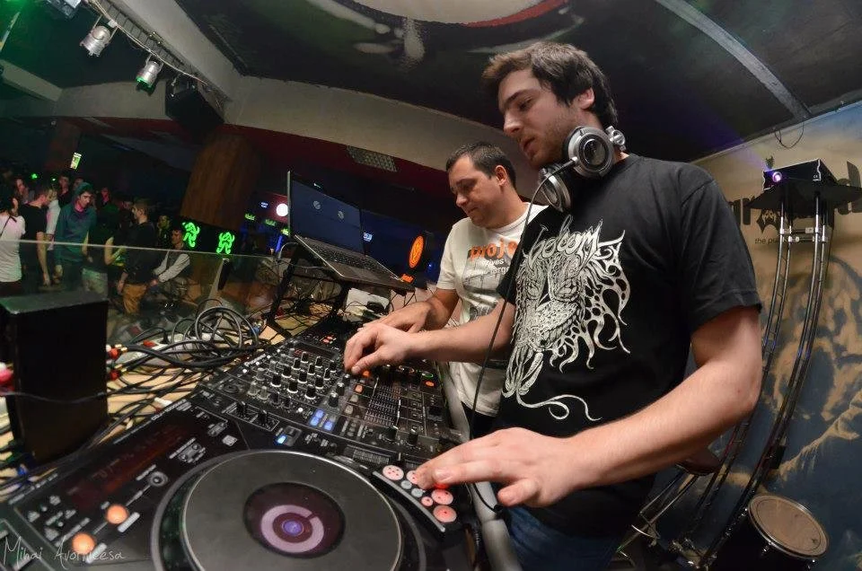 Two male DJs perform on a stage with a DJ controller and laptop, in front of a dance floor filled with people.