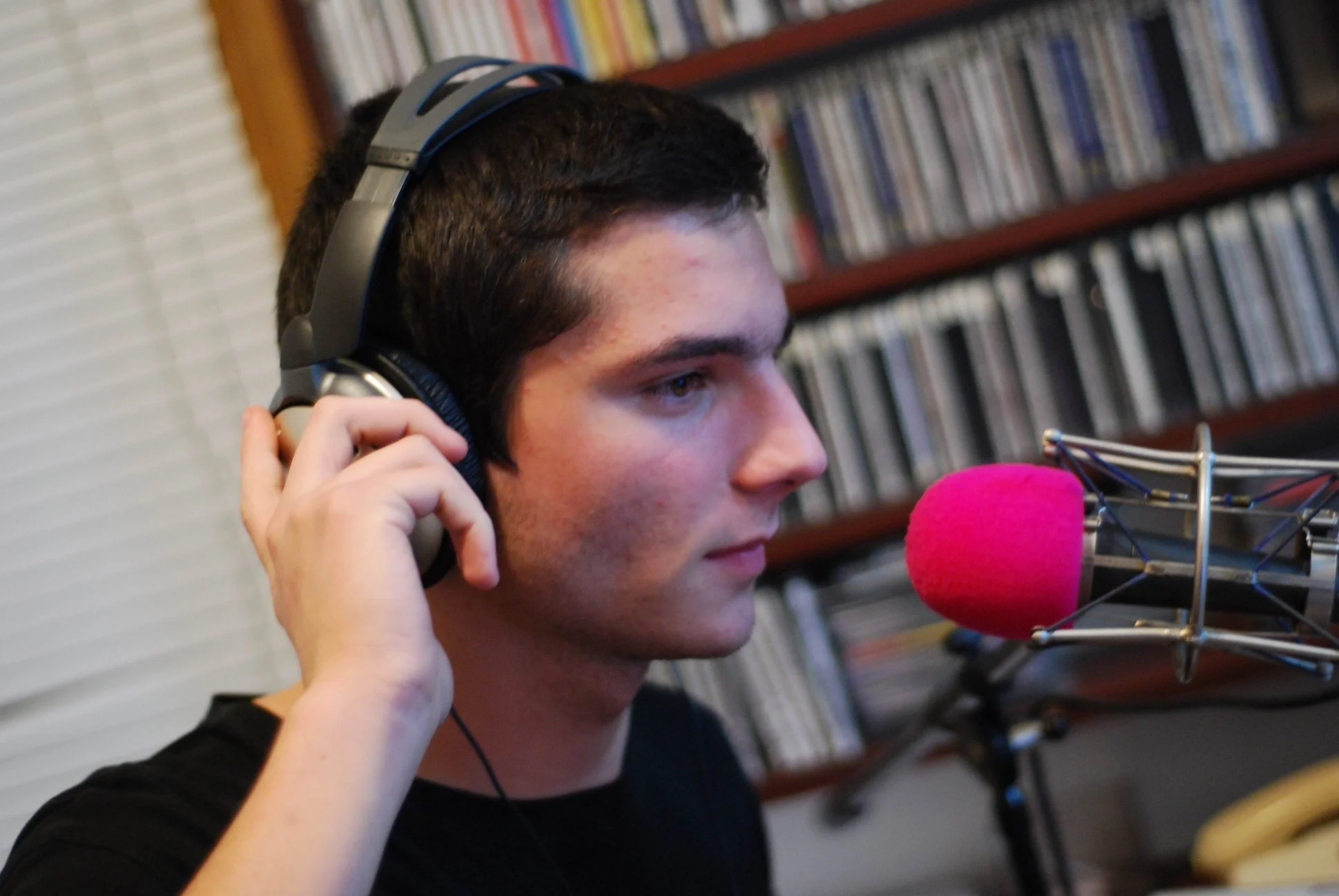 A young man with dark hair wearing headphones, speaking into a pink microphone in a recording studio with shelves of CDs or DVDs in the background.